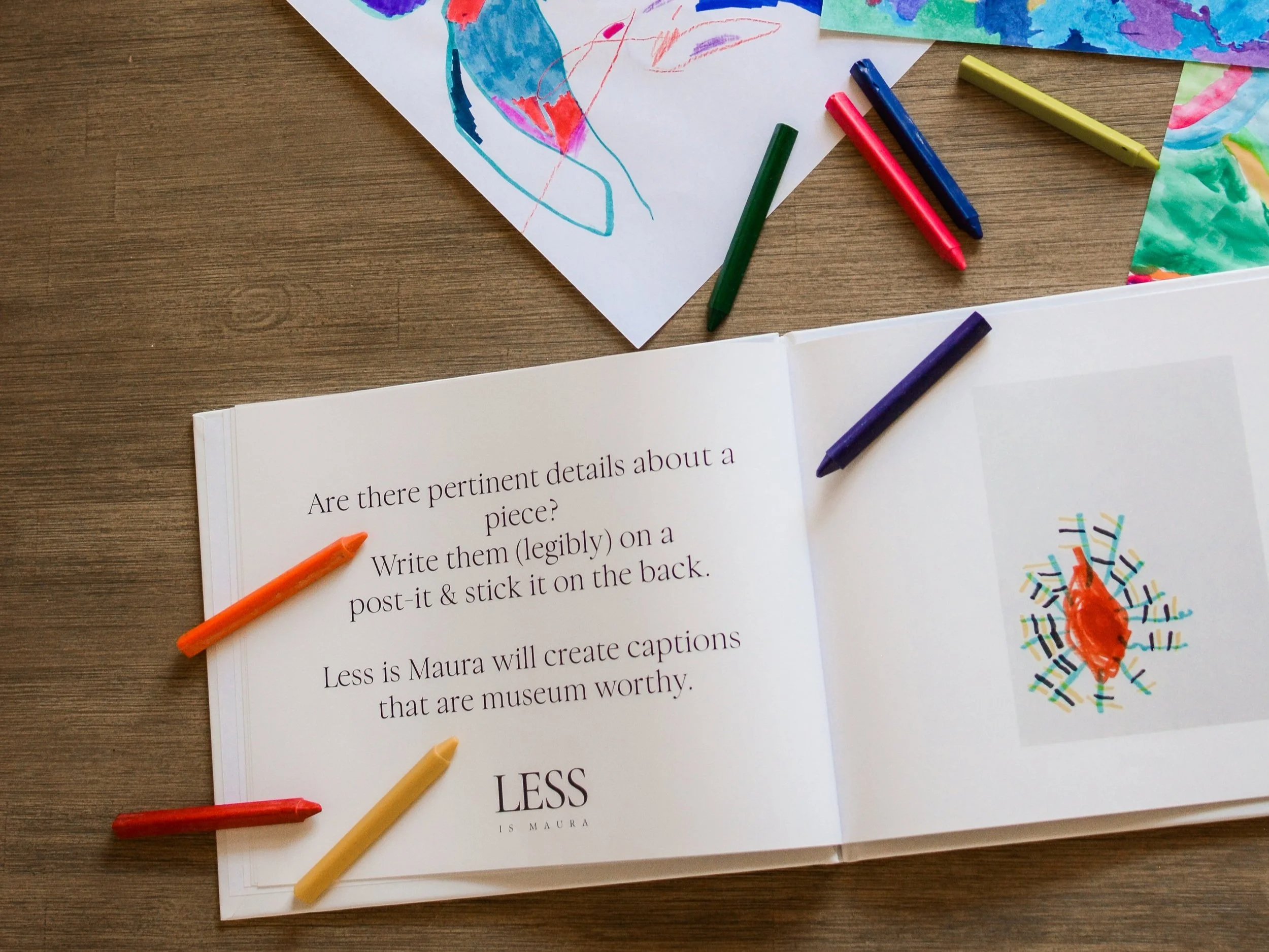 A photo album of children's art lies open with a description of an element of The Emerging Artist's Album service from Less is Maura. It lies open on a table with scattered crayons and pages of original artwork.