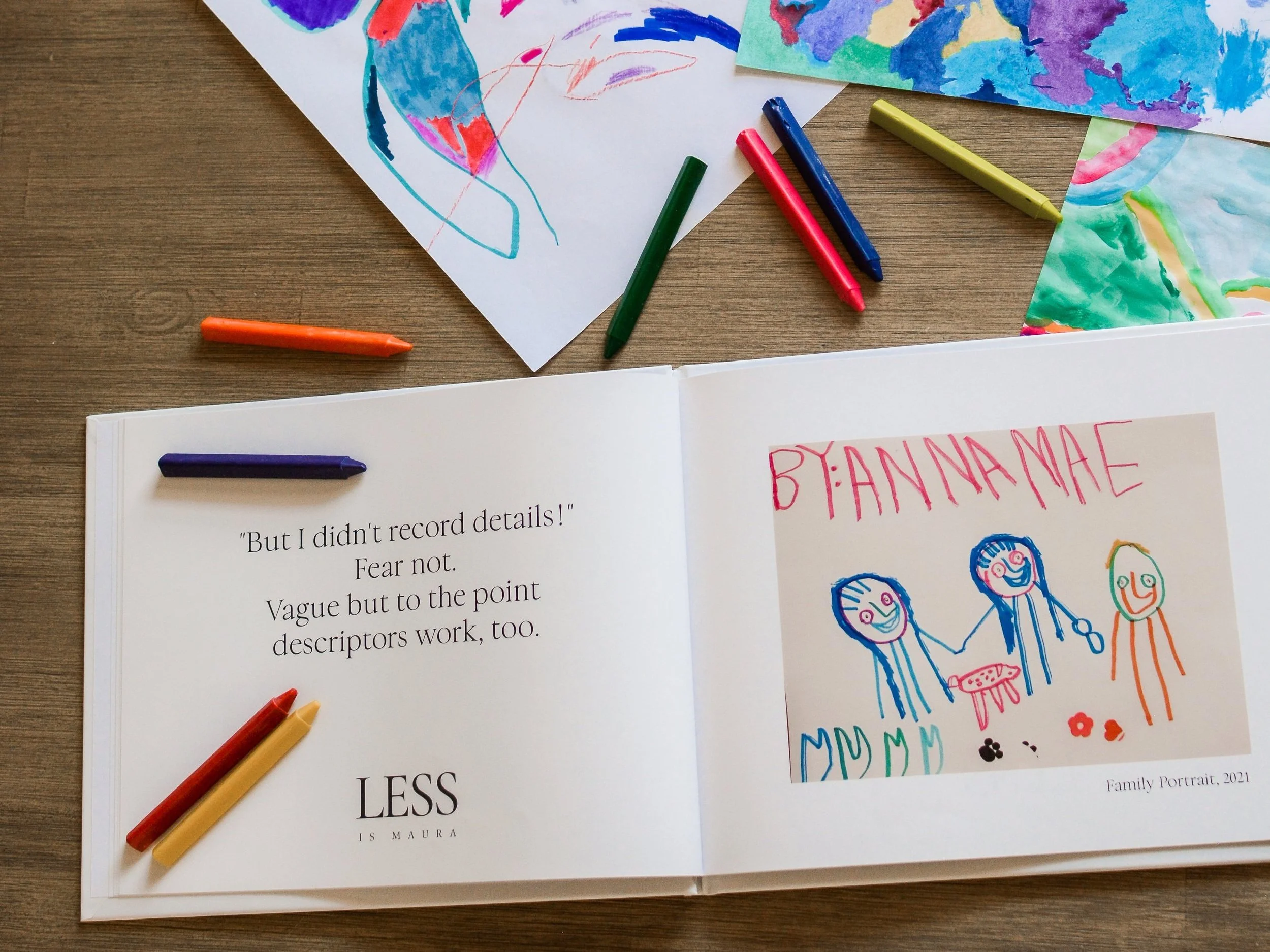 A photo album of children's art lies open with a description of an element of The Emerging Artist's Album service from Less is Maura. It lies open on a table with scattered crayons and pages of original artwork.