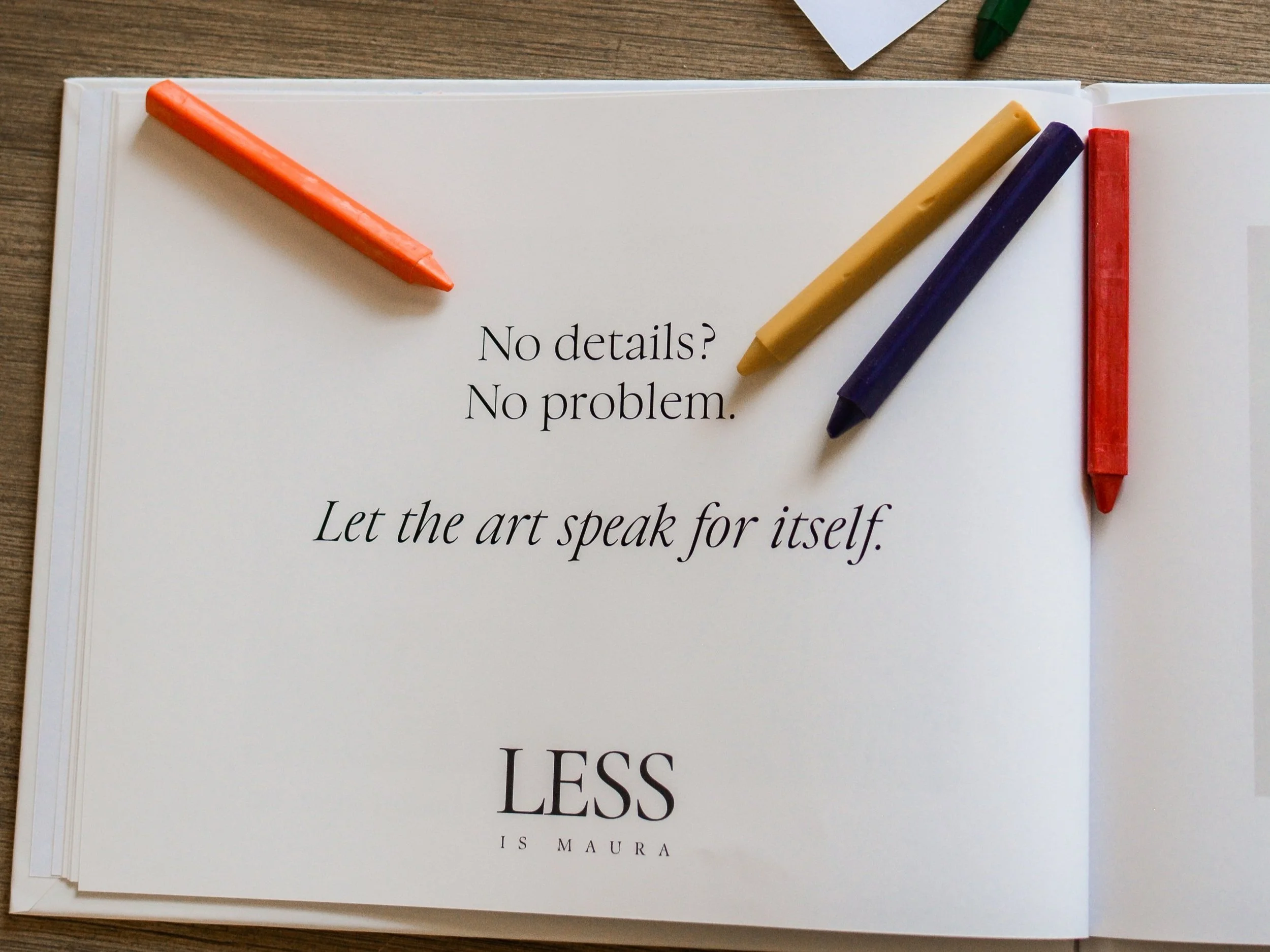 A photo album of children's art lies open with a description of an element of The Emerging Artist's Album service from Less is Maura. It lies open on a table with scattered crayons and pages of original artwork.