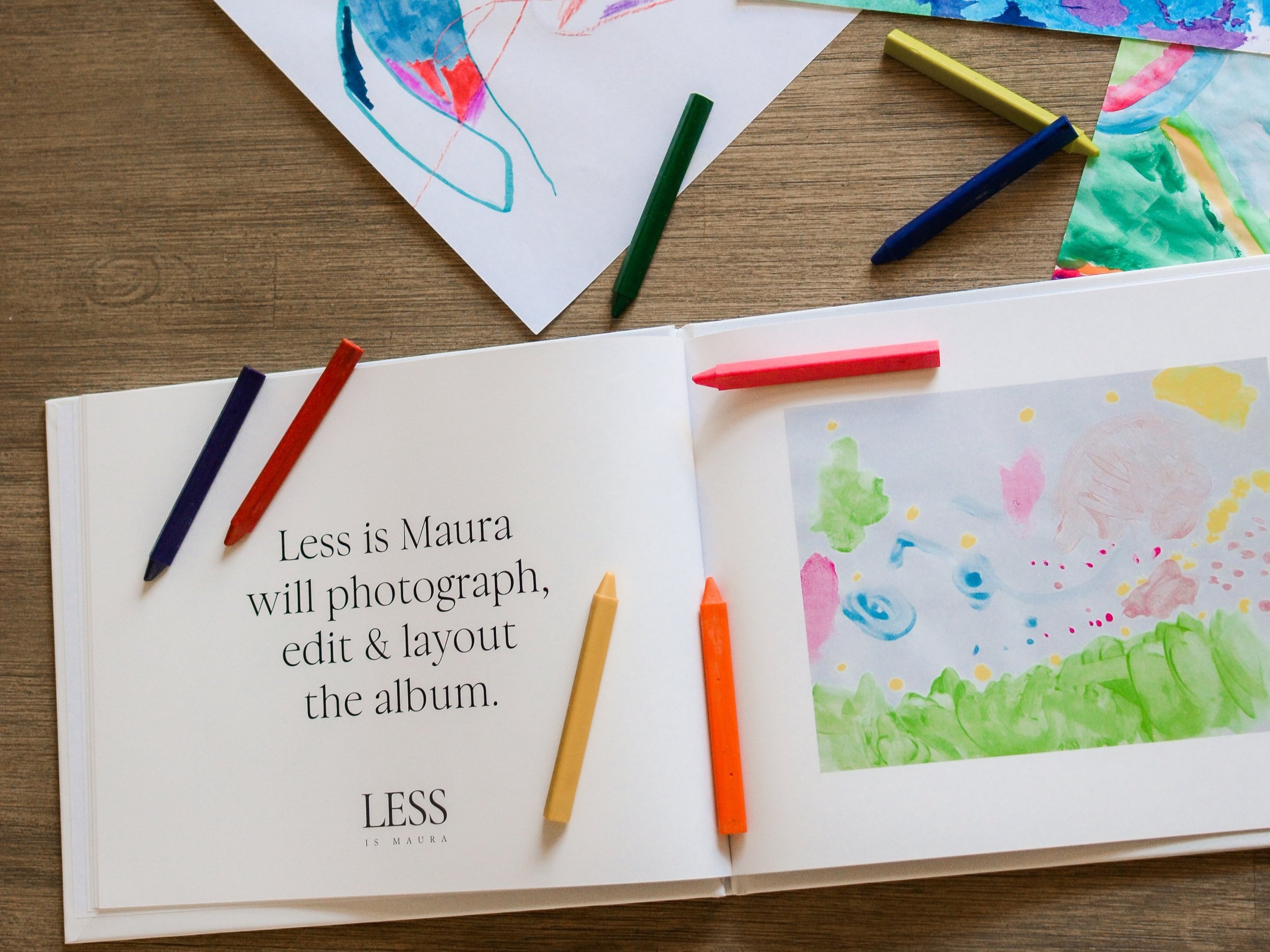 A photo album of children's art lies open with a description of an element of The Emerging Artist's Album service from Less is Maura. It lies open on a table with scattered crayons and pages of original artwork.