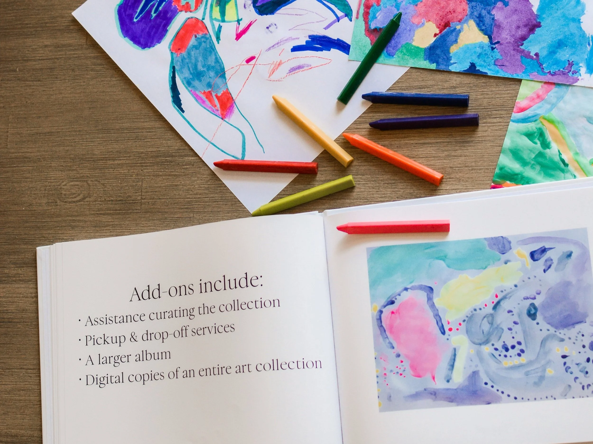 A photo album of children's art lies open with a description of an element of The Emerging Artist's Album service from Less is Maura. It lies open on a table with scattered crayons and pages of original artwork.
