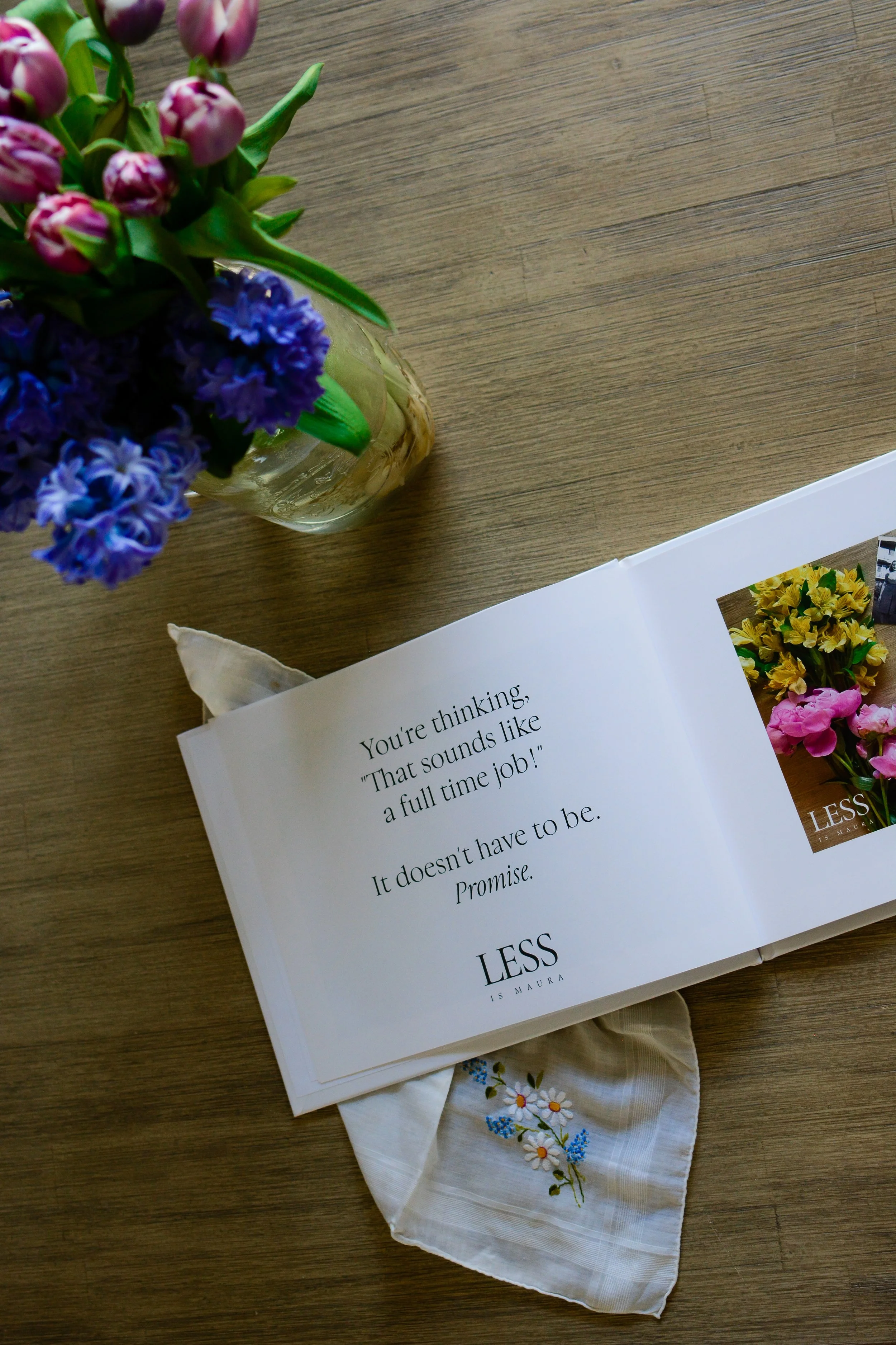 Looking down from above on a photo album with a picture and a description of instructions, lying open on a table with a vase of flowers and a handkerchief.