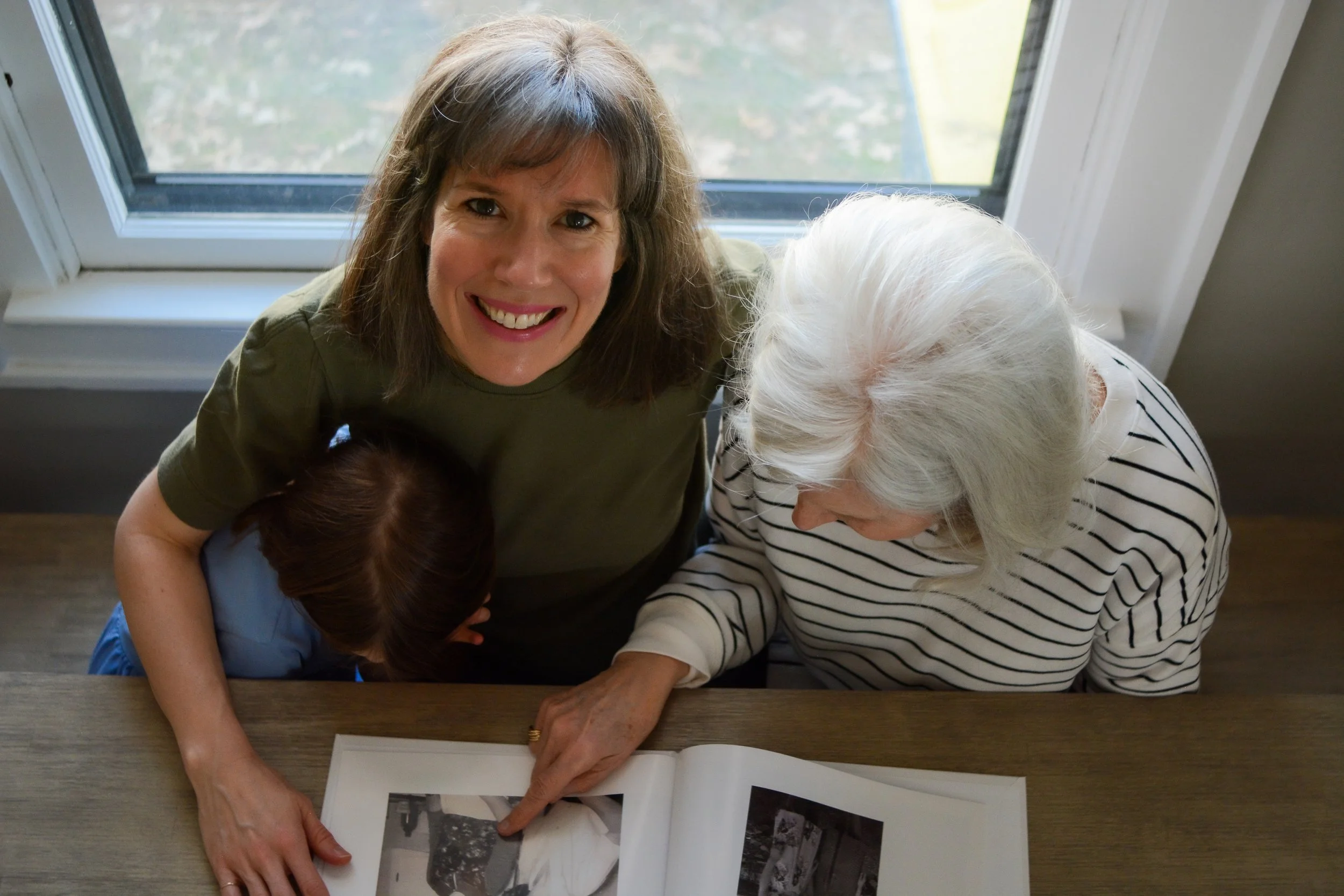 A mother looks up to the camera, her arms around her seven year old daughter and her mom - a white haired grandma - as they look through a photo album, The Grandparent Album.