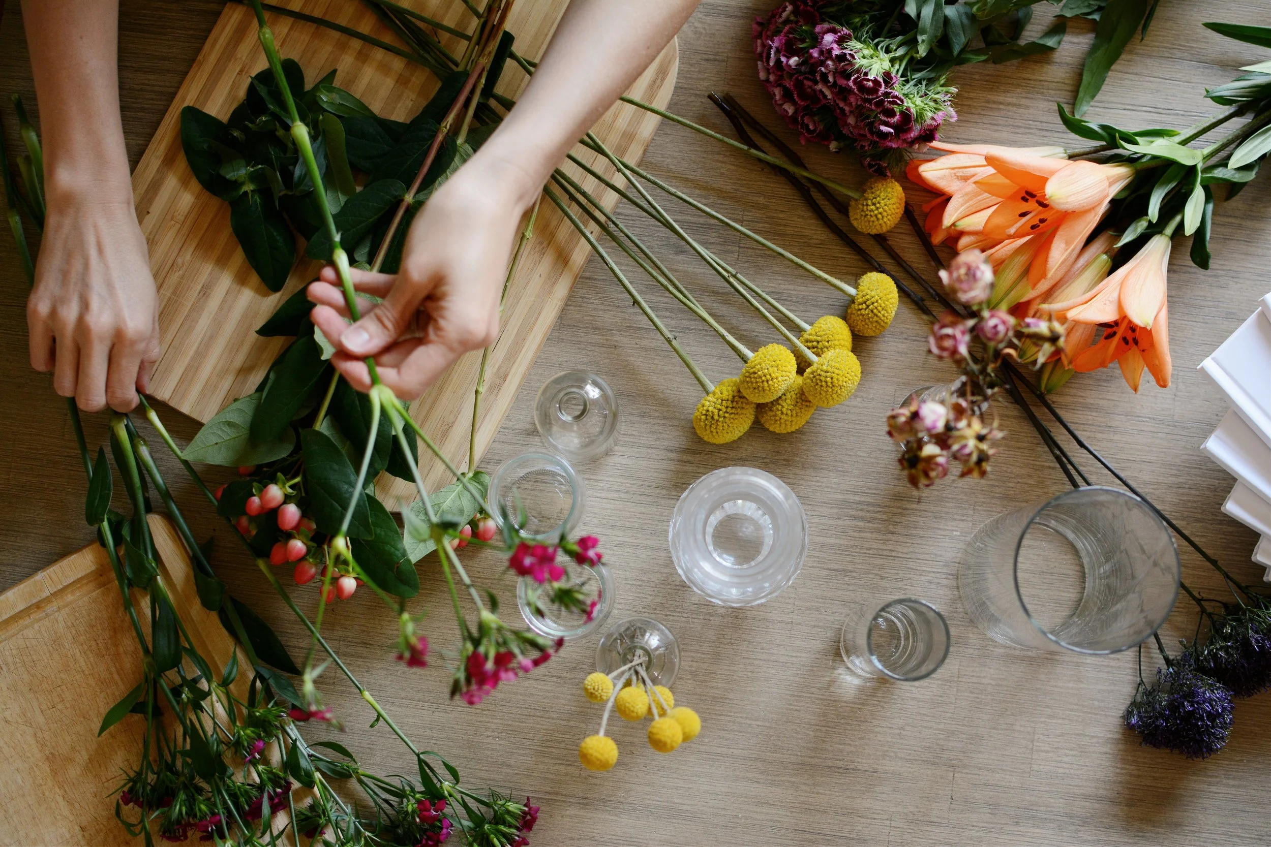 Flowers and vases lay scattered across a table. A pair of hands arranges the flowers.