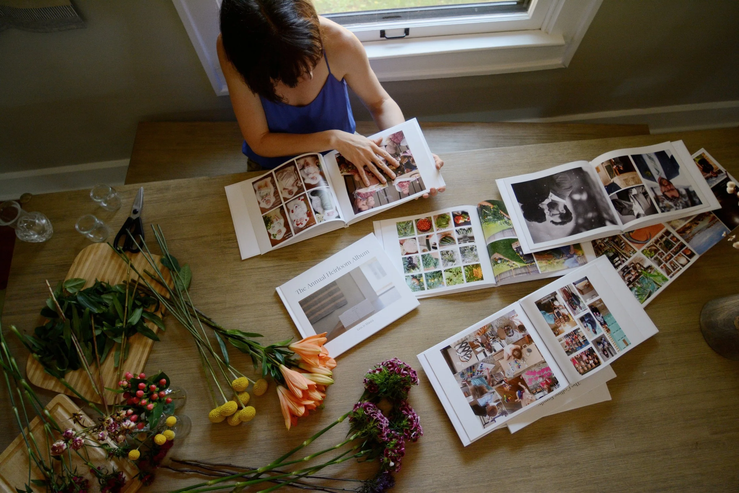 Looking from above on a woman flipping through a photo albums, with various other albums opened and scattered around the table alongside various flowers waiting to be turned into bouquets.