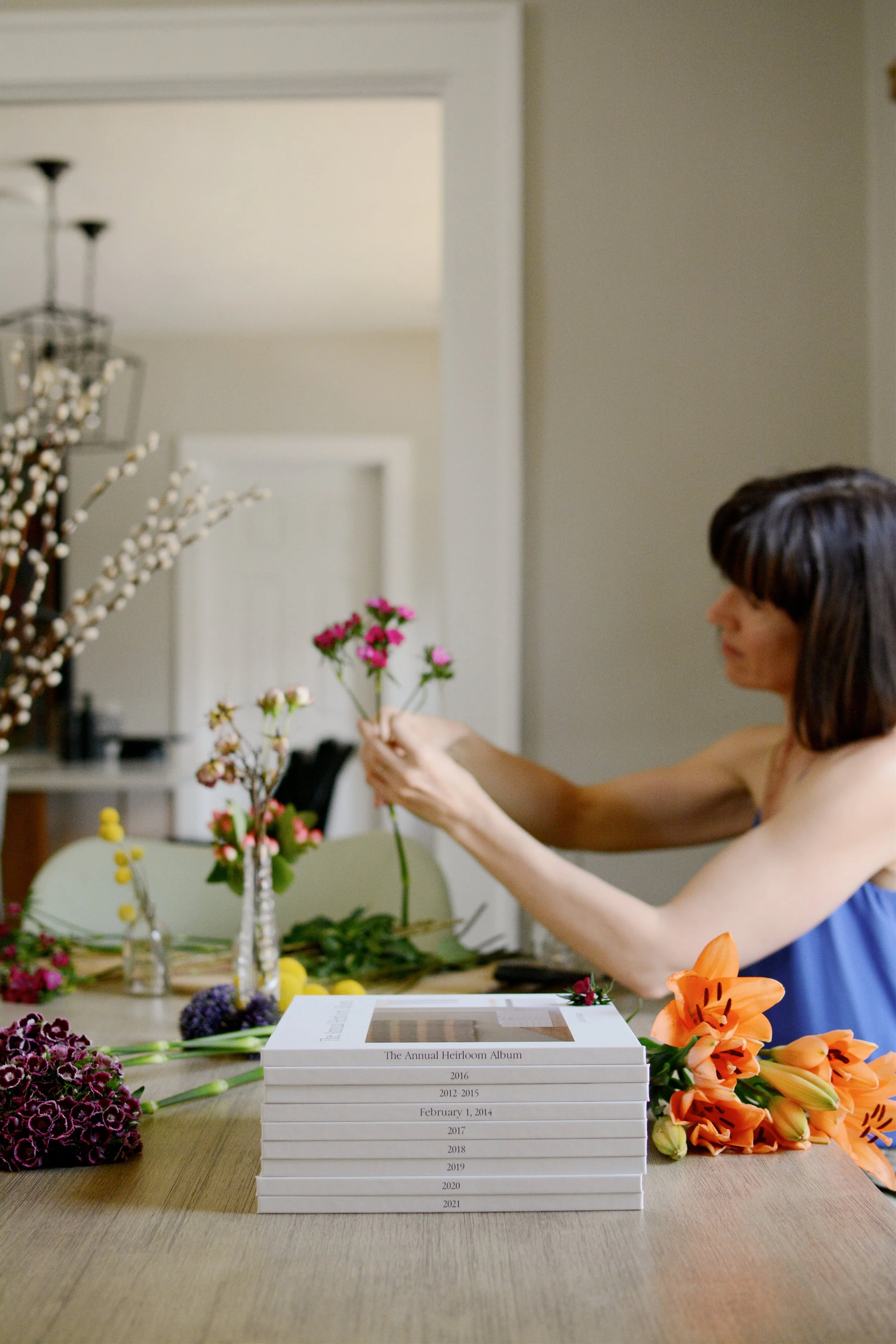 A woman arranges flowers in the background, a stack of albums sits on the table in the foreground.