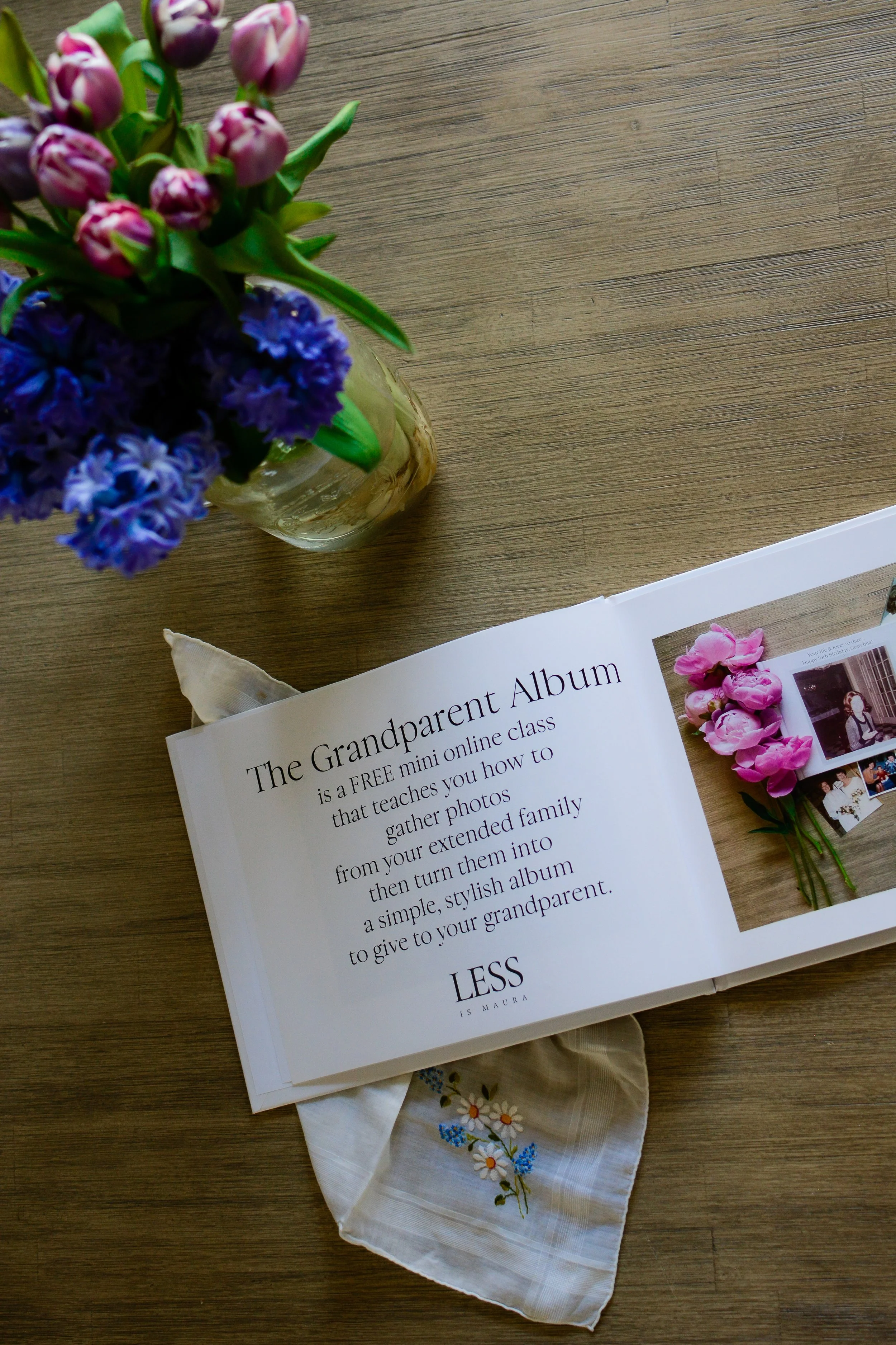 Looking down from above on a photo album, lying open on a table with a vase of flowers and a handkerchief.