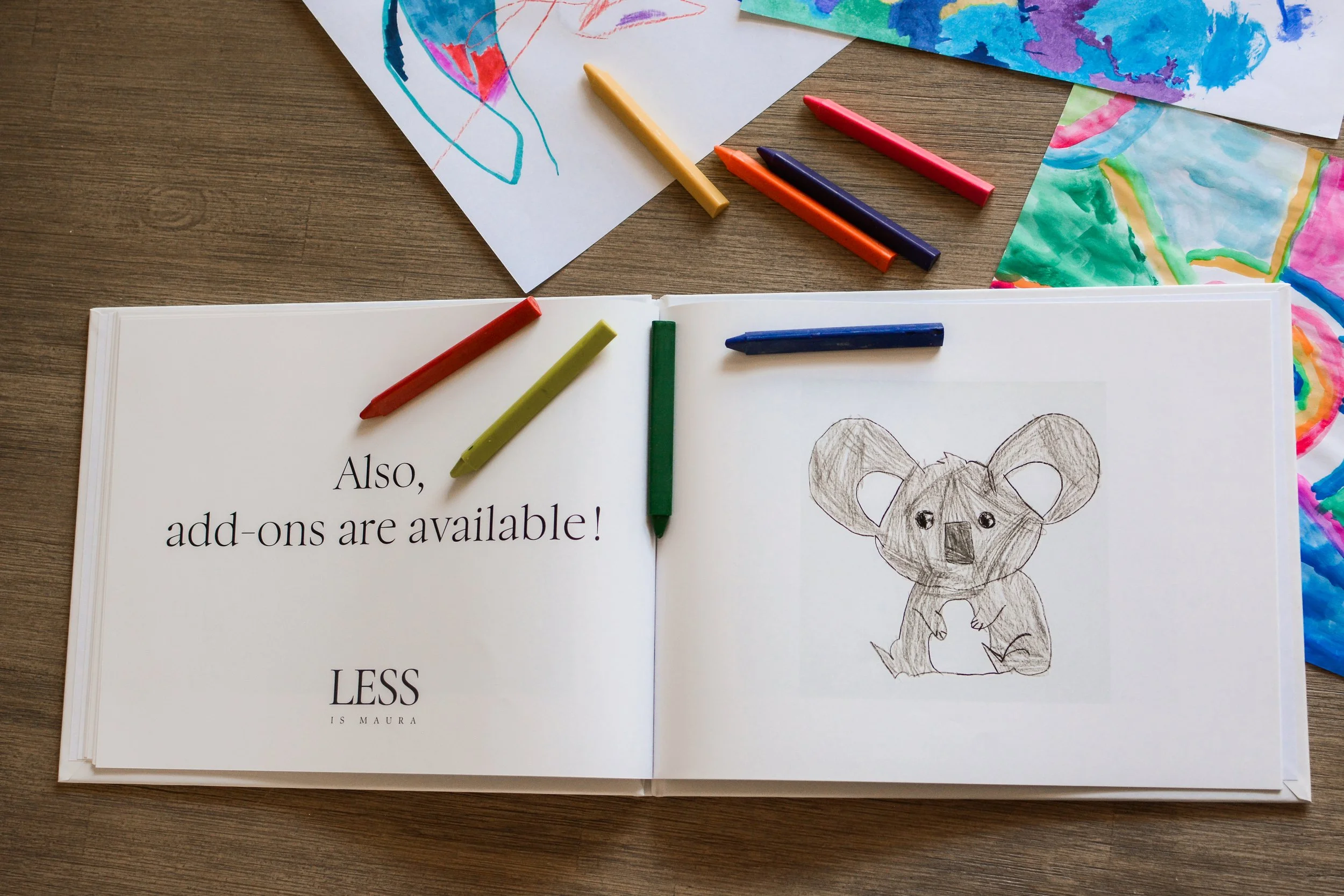 A photo album of children's art lies open with a description of an element of The Emerging Artist's Album service from Less is Maura. It lies open on a table with scattered crayons and pages of original artwork.