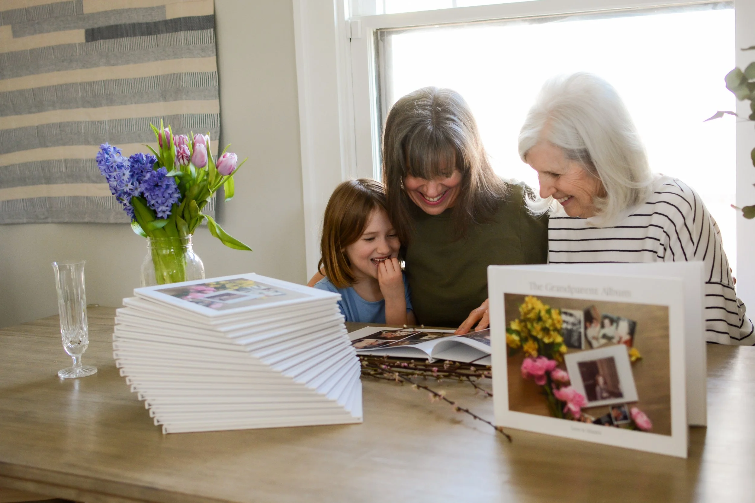 A seven yar old daughter with her mom and grandma look through a photo album in the background, with another photo album entitled "The Grandparent Album" standing up in the foreground.