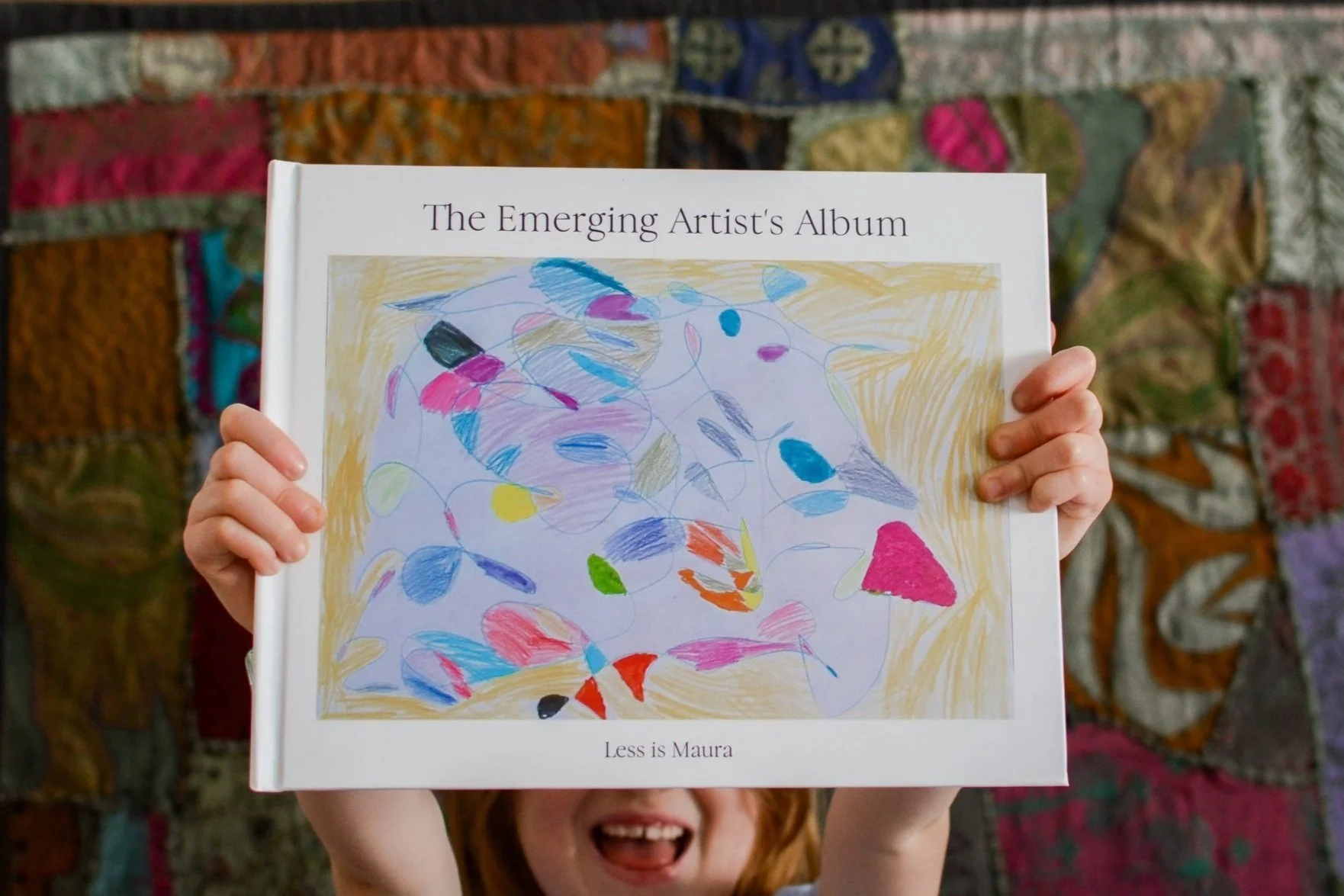 A little girl smiles as she holds an art album in front of her face.