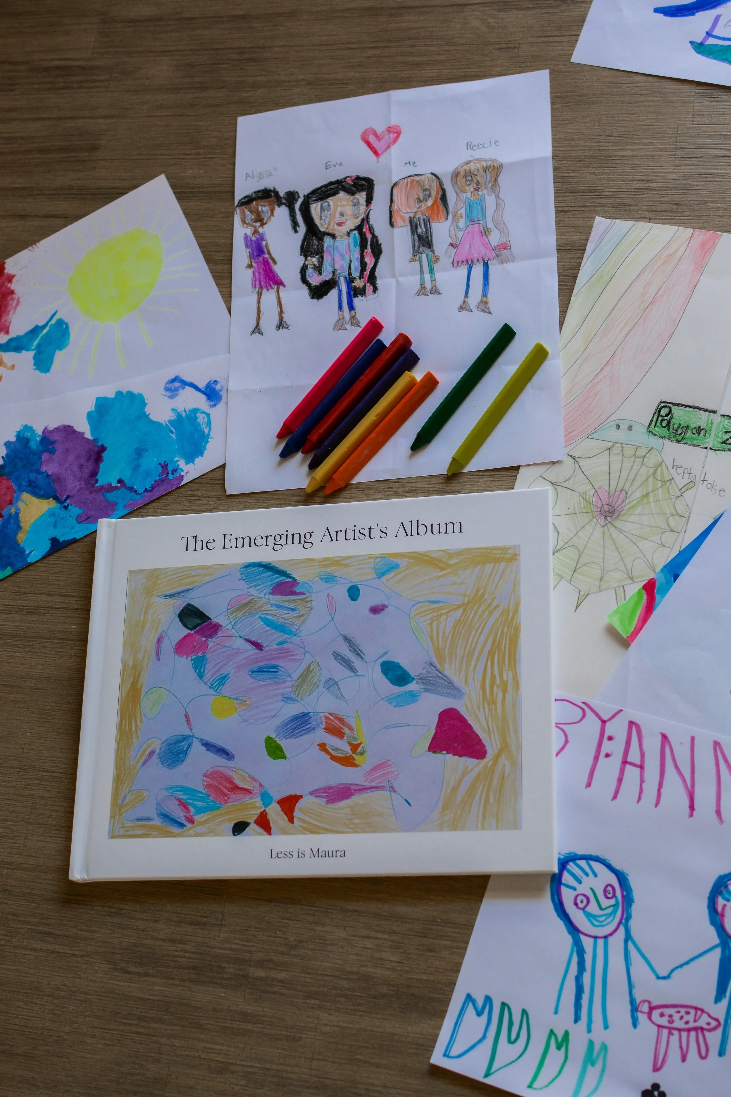 A kid art album lays on a table with kid artwork and crayons scattered all around it.