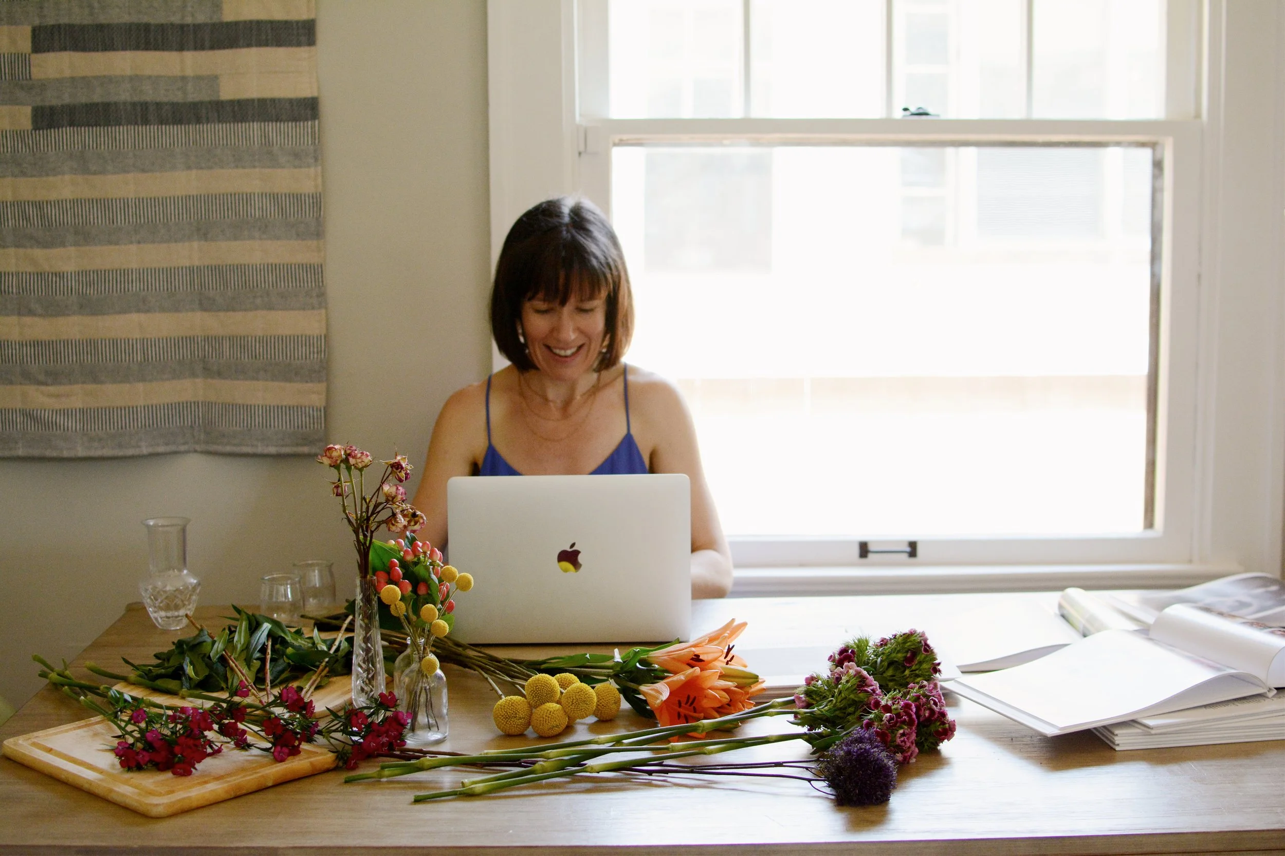 A woman sits at a table and types o na laptop, flowers scattered on the table in front of the laptop.