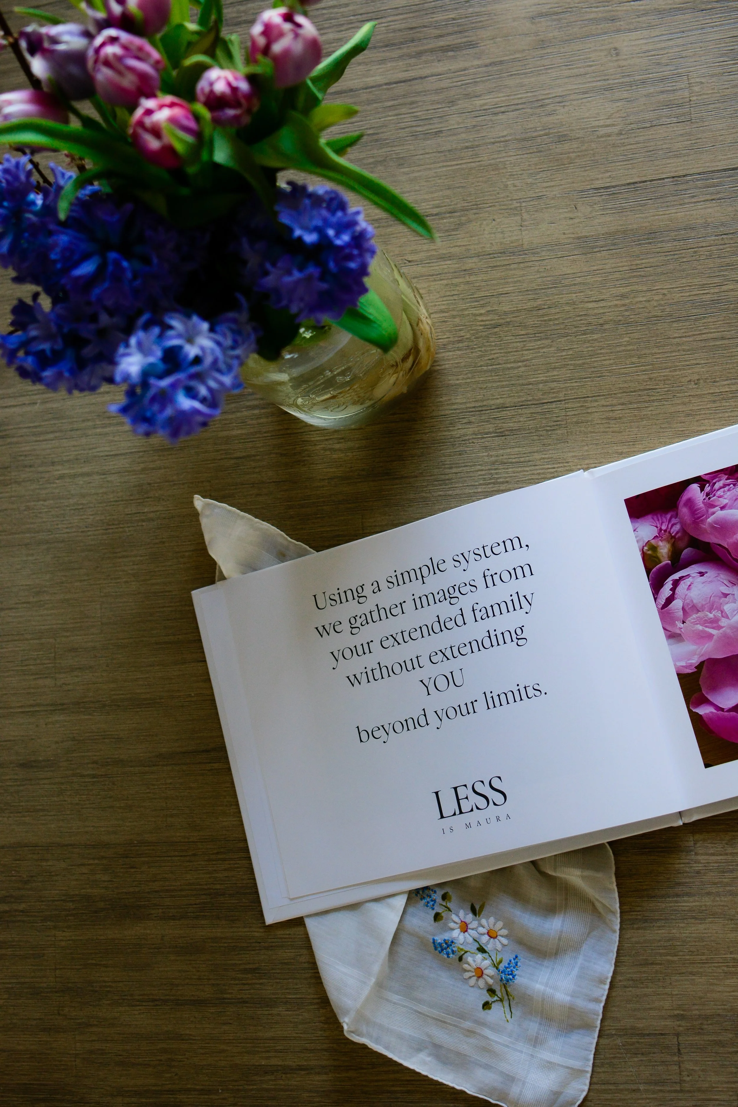 Looking down from above on a photo album with a picture and a description of instructions, lying open on a table with a vase of flowers and a handkerchief.