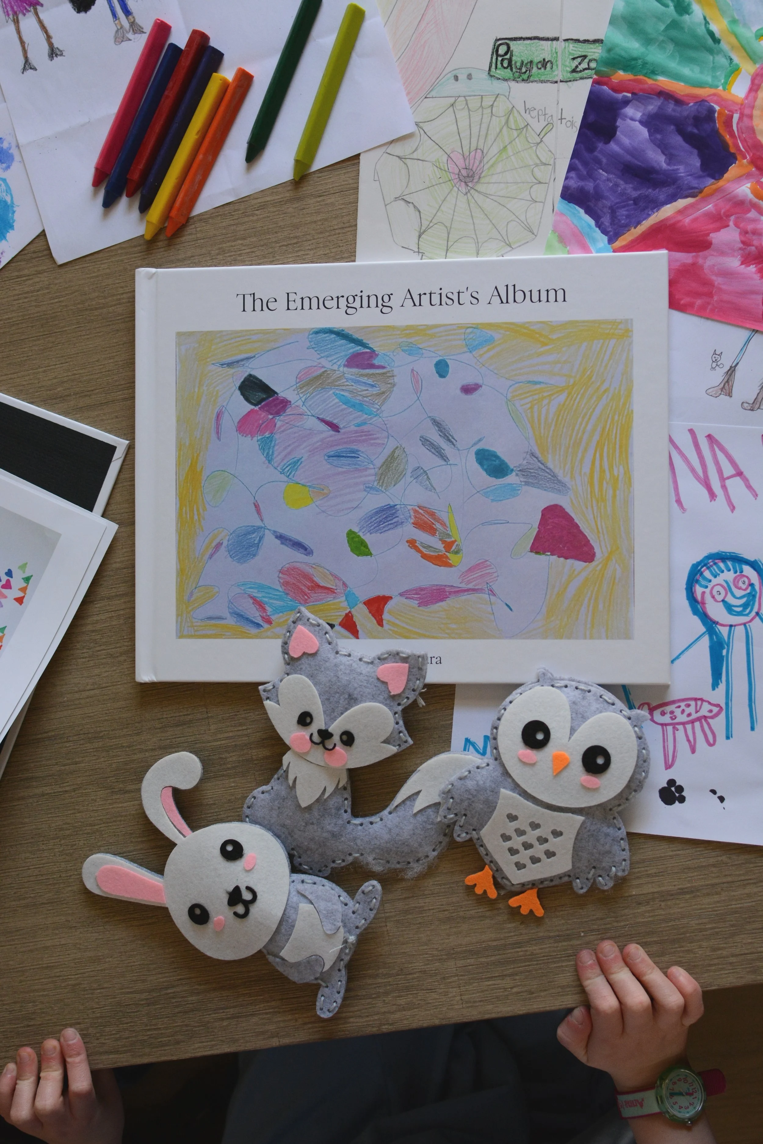 A kid art album lays on a table, kid art and crayons and kid-made stuffed animals laying all around it, with two little hands at the edge of the table.