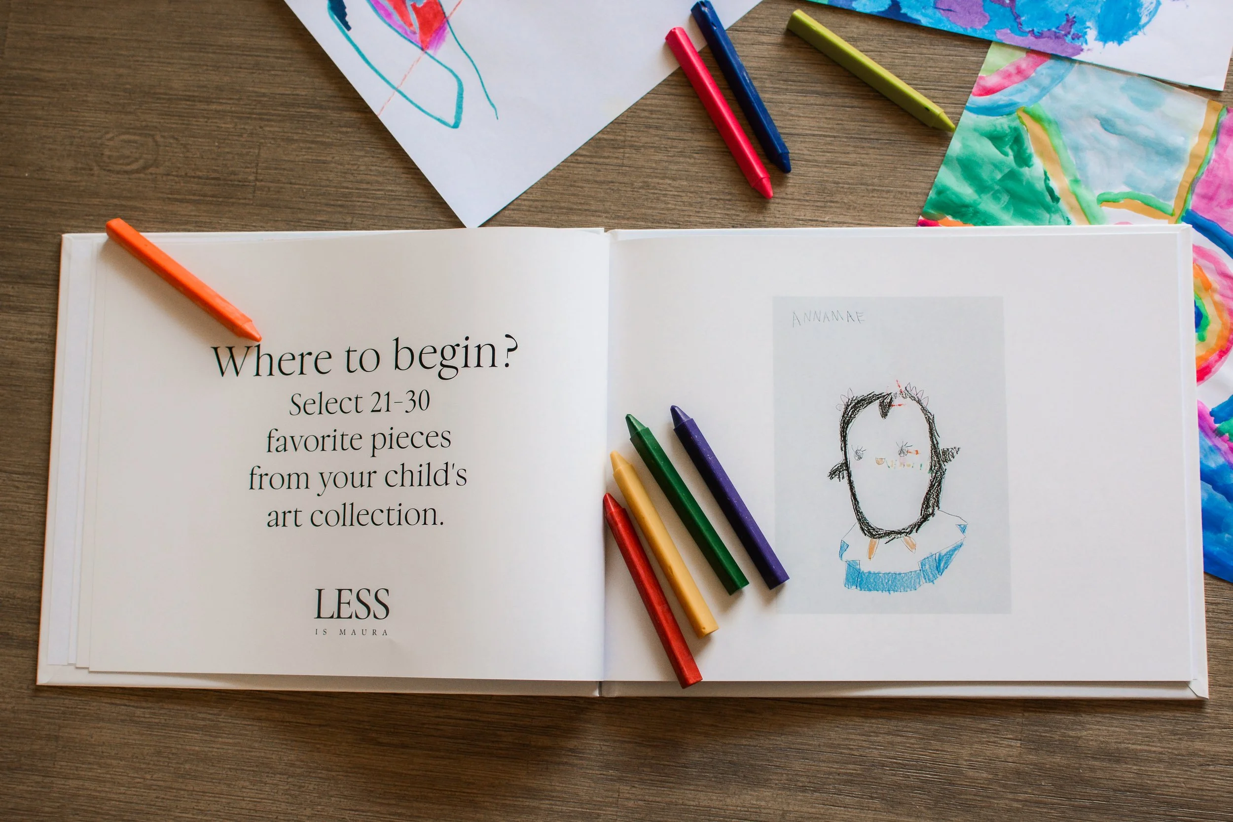 A photo album of children's art lies open with a description of an element of The Emerging Artist's Album service from Less is Maura. It lies open on a table with scattered crayons and pages of original artwork.