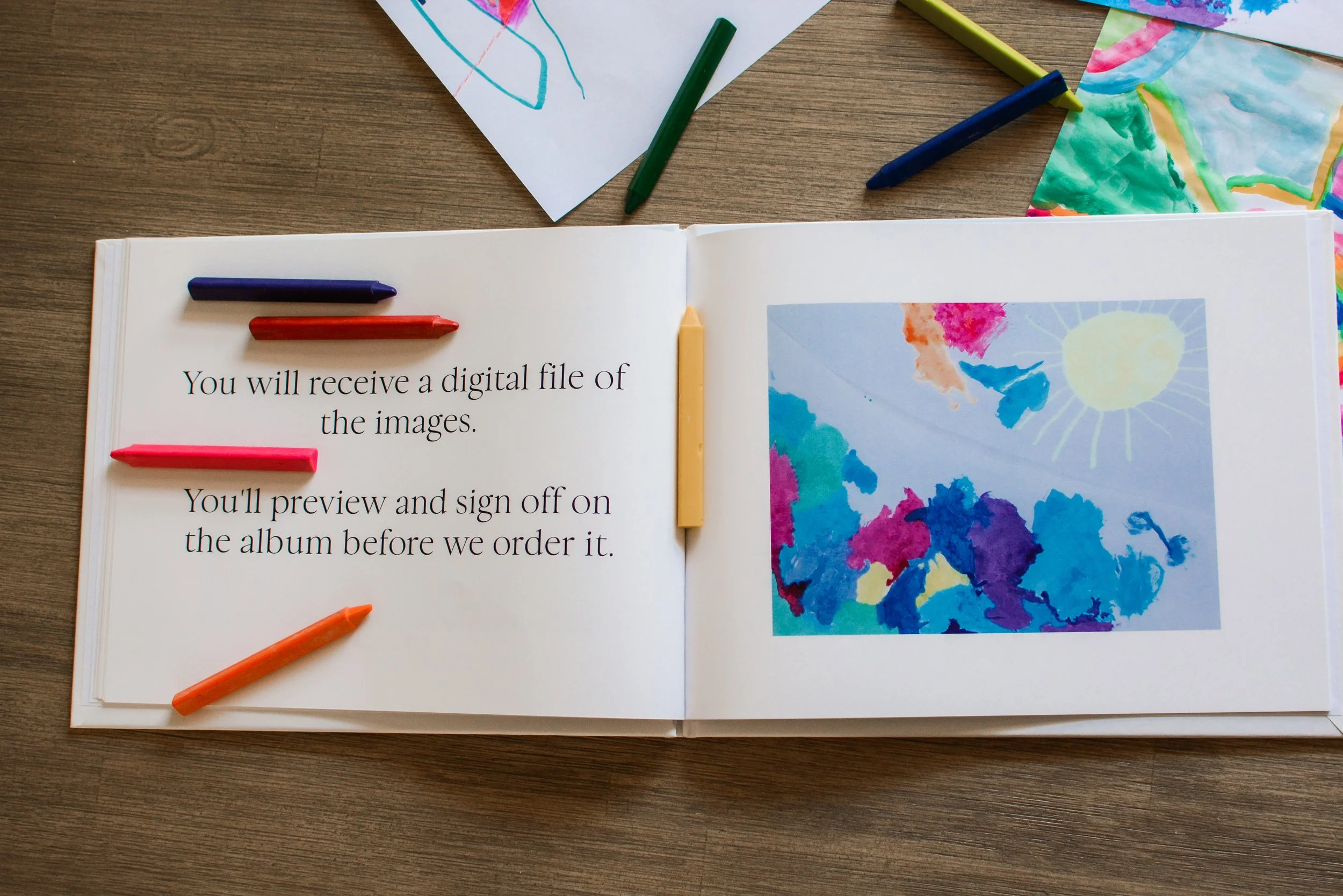 A photo album of children's art lies open with a description of an element of The Emerging Artist's Album service from Less is Maura. It lies open on a table with scattered crayons and pages of original artwork.