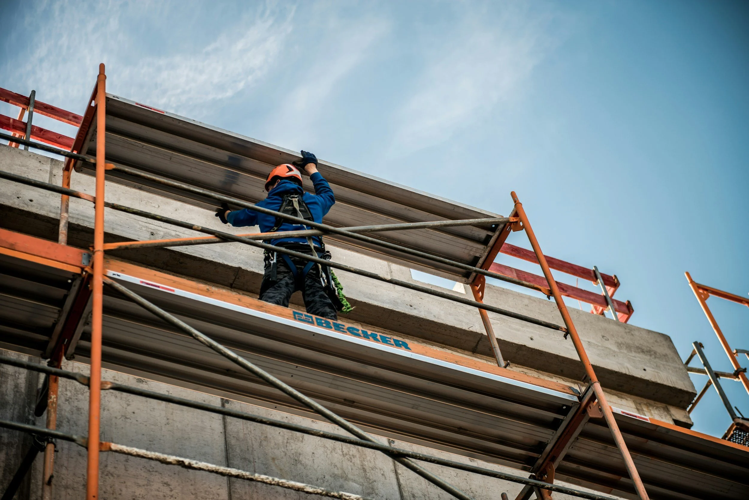 A construction worker wearing a helmet and harness is standing on a scaffold while working on a building's concrete structure against a partly cloudy sky.