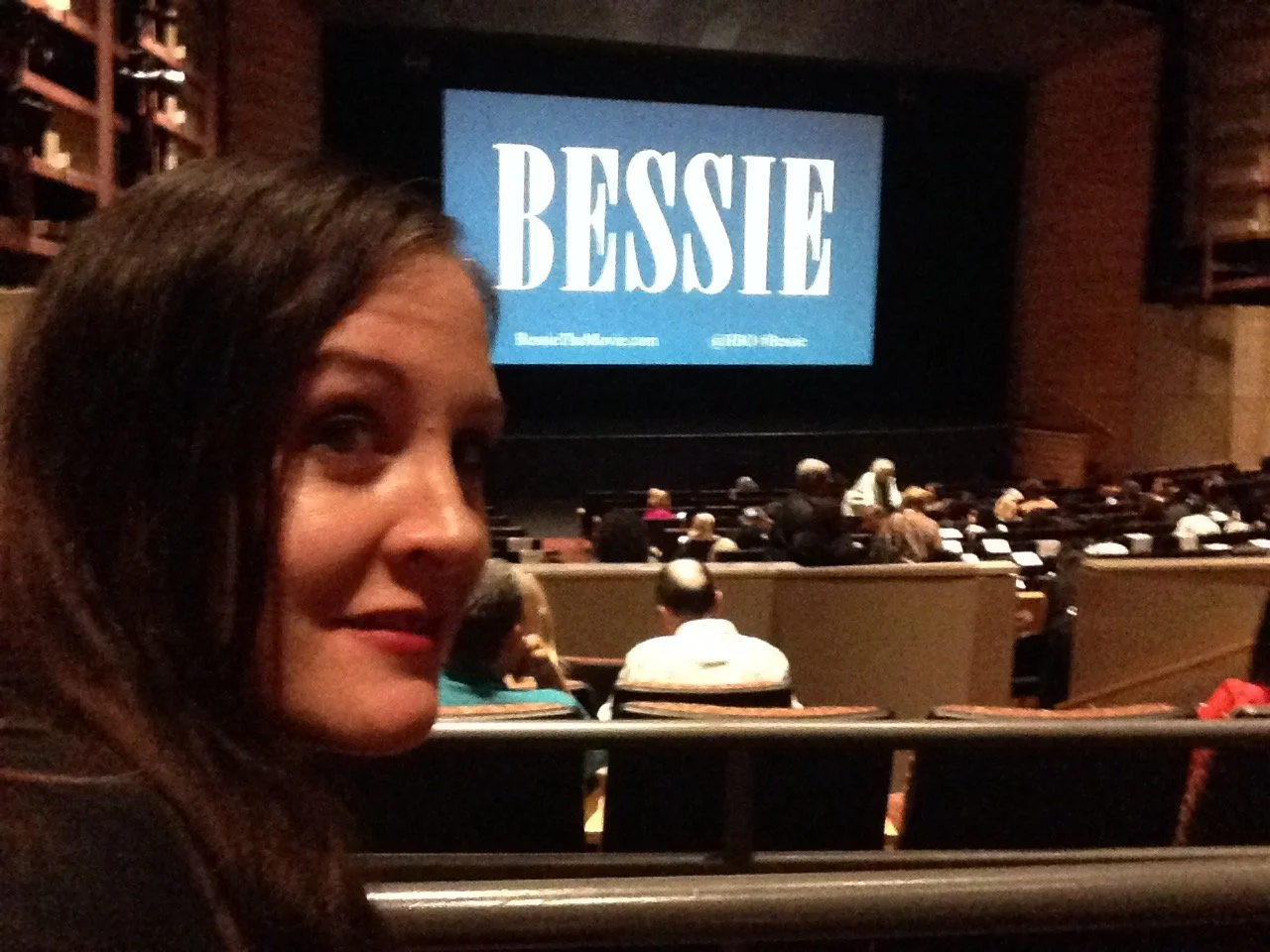 A woman with dark hair inside a theater, with a large screen in the background displaying the name 'BESSIE' with a blue background and white text. The theater is filled with people seating and conversing.