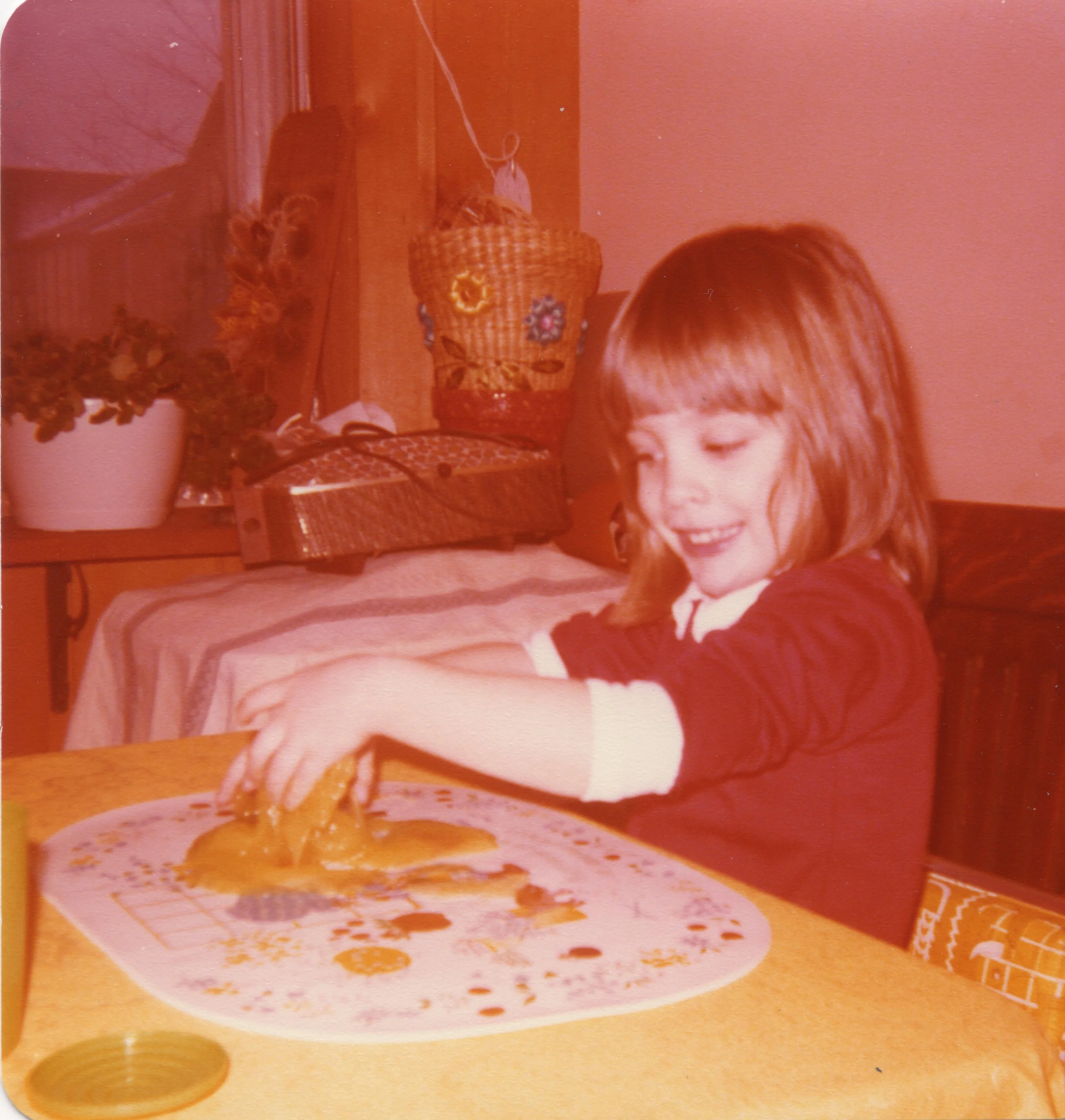 A child playing with paint at a table with decorative placemat, surrounded by a potted plant, woven basket, and vintage radio. The room has warm lighting and retro decor.