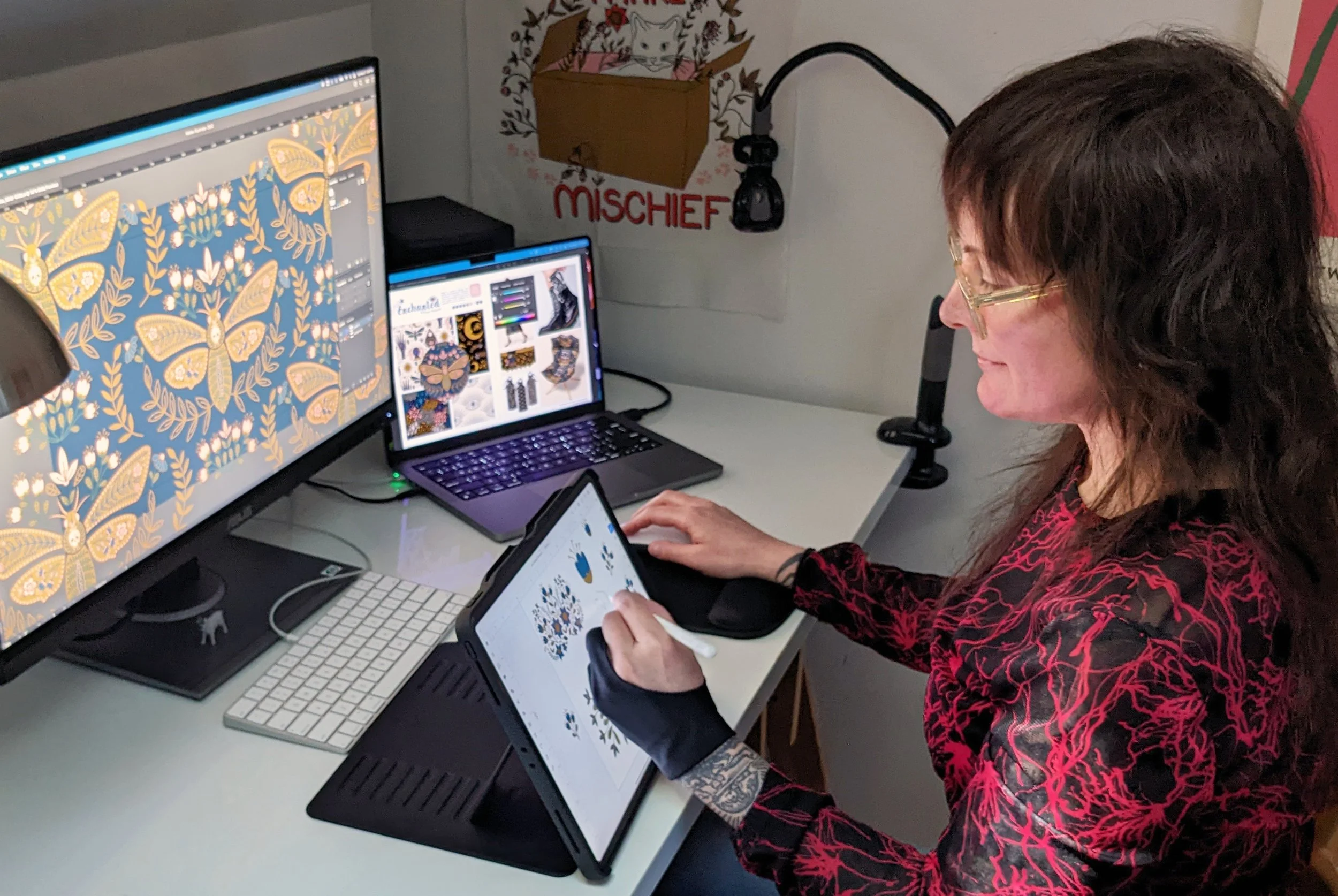 A woman working at a desk with multiple screens, creating butterfly and floral digital art. She is using a stylus on a drawing tablet, with a tattooed arm visible.