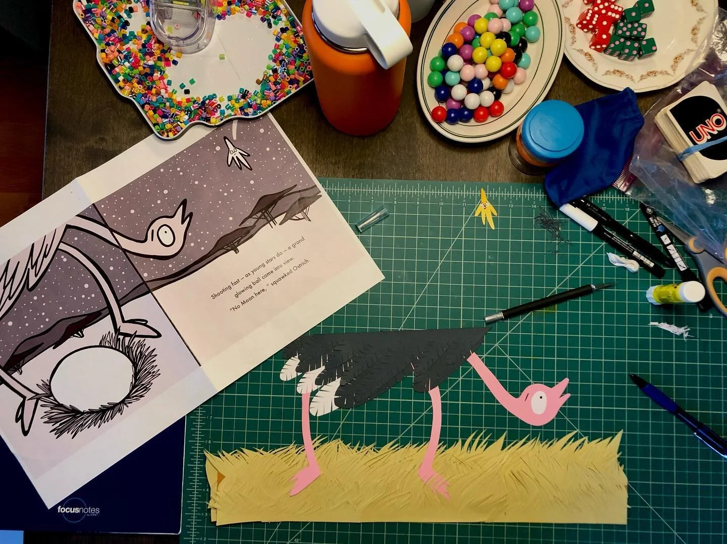 I&rsquo;m currently at my parents cabin cutting up a piece for the picture book I&rsquo;m working on. To the left is the book dummy I drew that is used as a template for the final art. Making a paper ostrich surrounded by tiny people toy chaos is 👌.