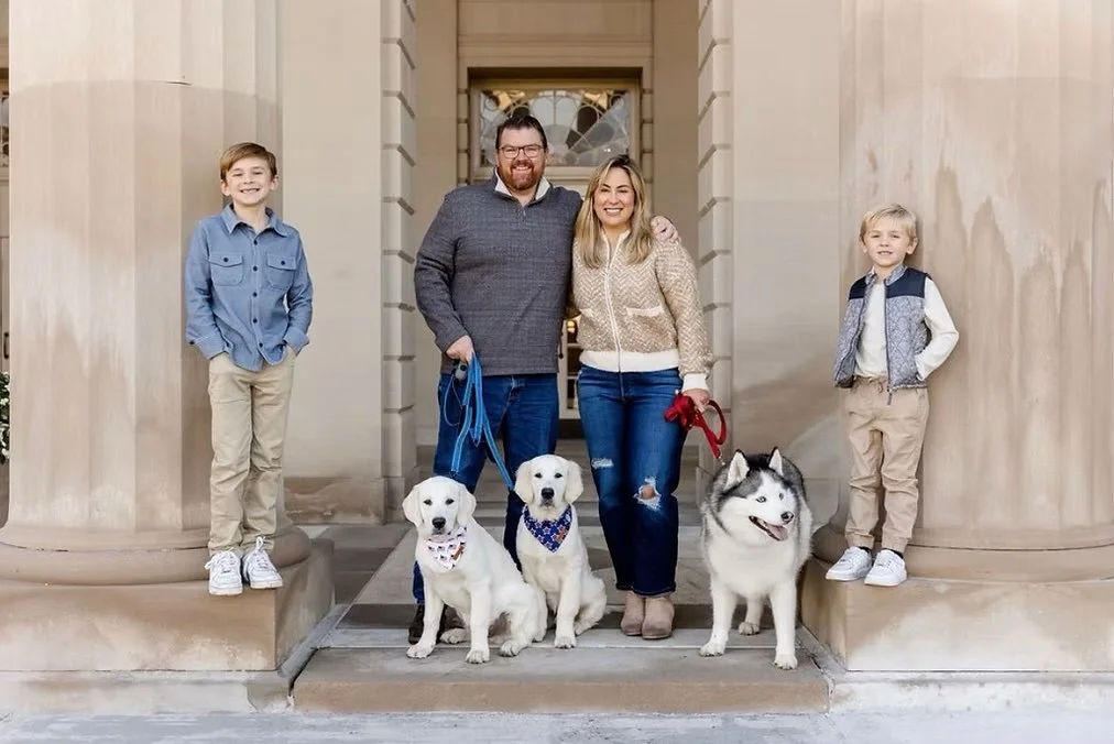 A family of four standing on the steps of a building with two boys, a man, and a woman, along with three dogs, including a husky, a husky puppy, and two lab retrievers. They are smiling and posing for the photo.