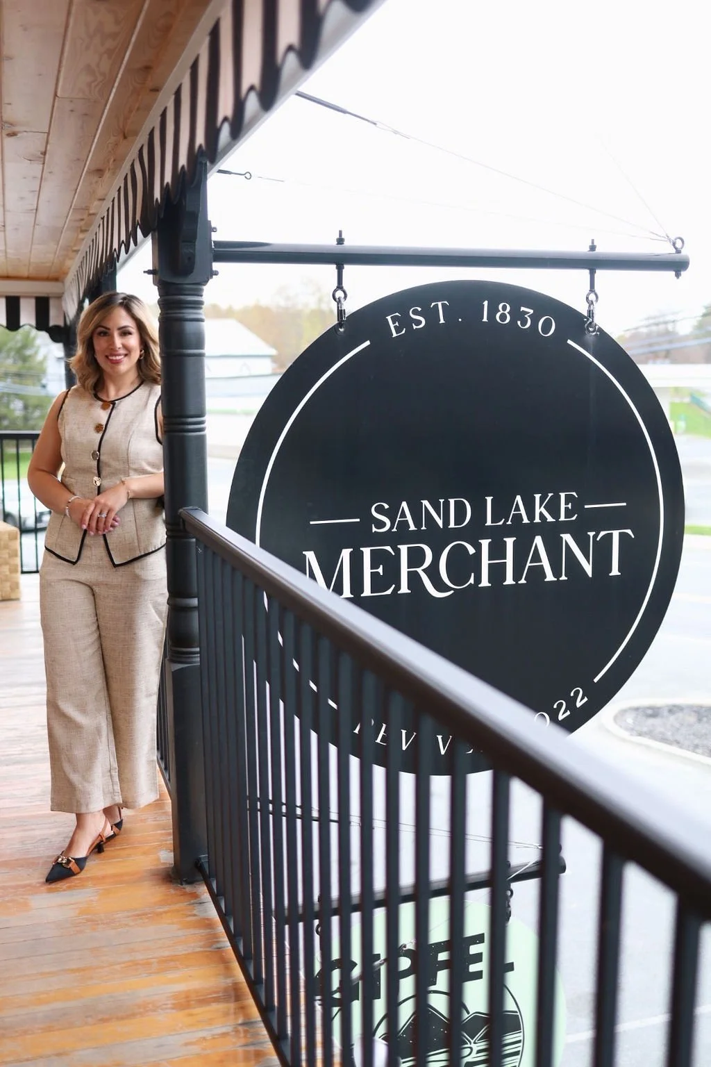 A woman in beige business attire standing on a wooden porch with a black railing, smiling near a large circular sign for Sand Lake Merchant, established 1830, with overcast skies in the background.