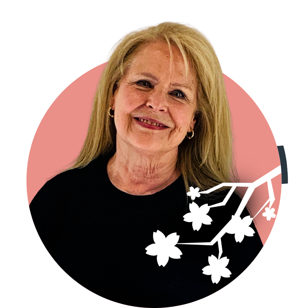 Thumbnail image of associate therapist Michelle Stockwell: A smiling elderly woman with blonde hair and earrings, wearing a black top, with a graphic of white cherry blossoms and pink, purple, and dark teal geometric shapes in the background.