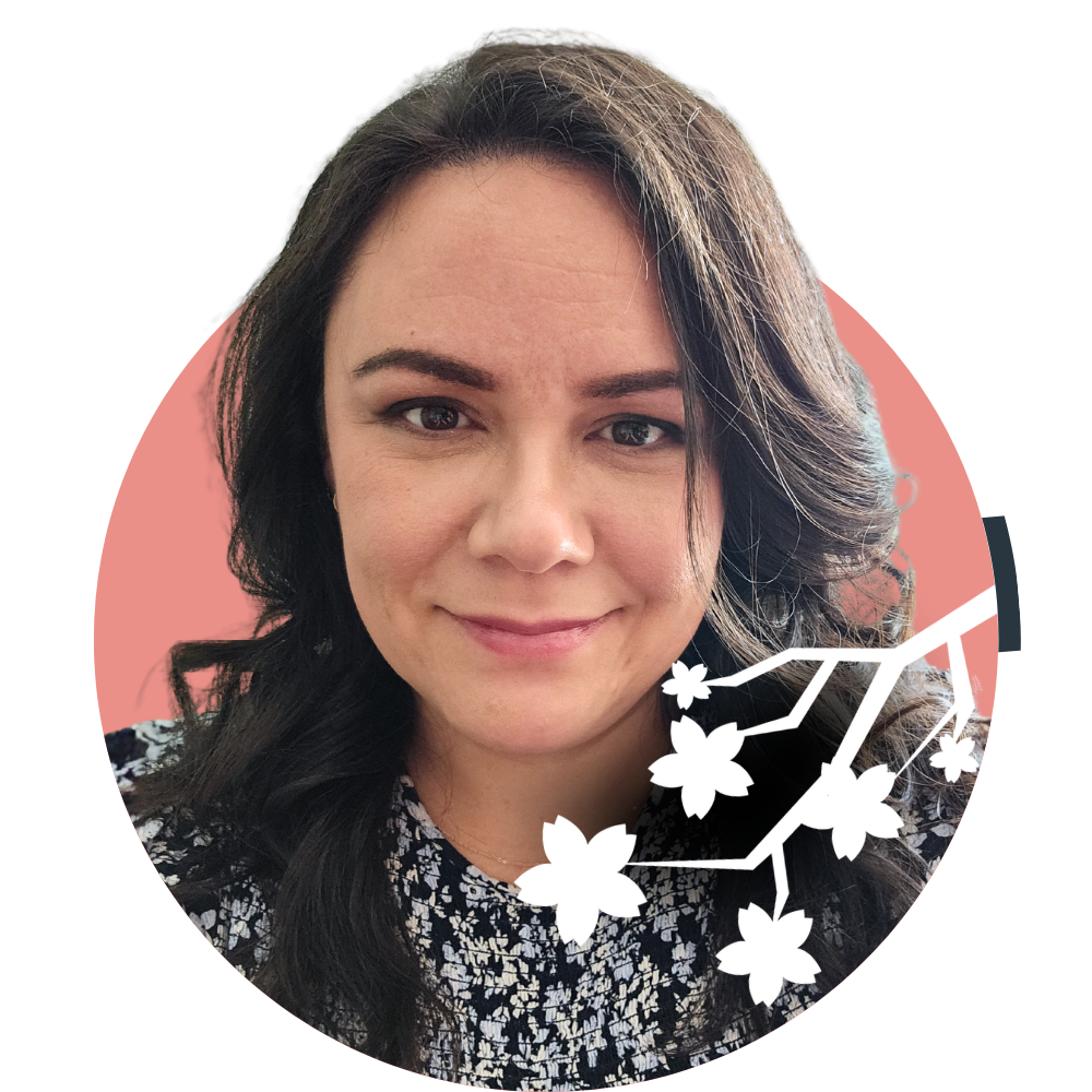Thumbnail image of associate therapist Amy Commanda: A woman with dark brown, wavy hair and light skin smiling at the camera, wearing a patterned top. An illustration of cherry blossoms and branches is overlaid on the bottom right of the photo.
