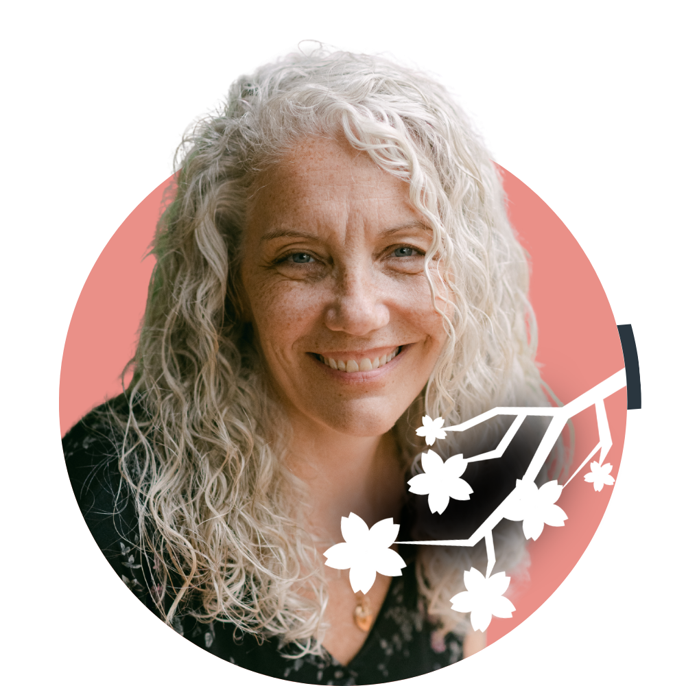 Thumbnail image of associate therapist Beth Lewis: A woman with long, curly gray hair smiling at the camera, wearing a black floral top, with a pink background and white flower and branch graphic overlay.