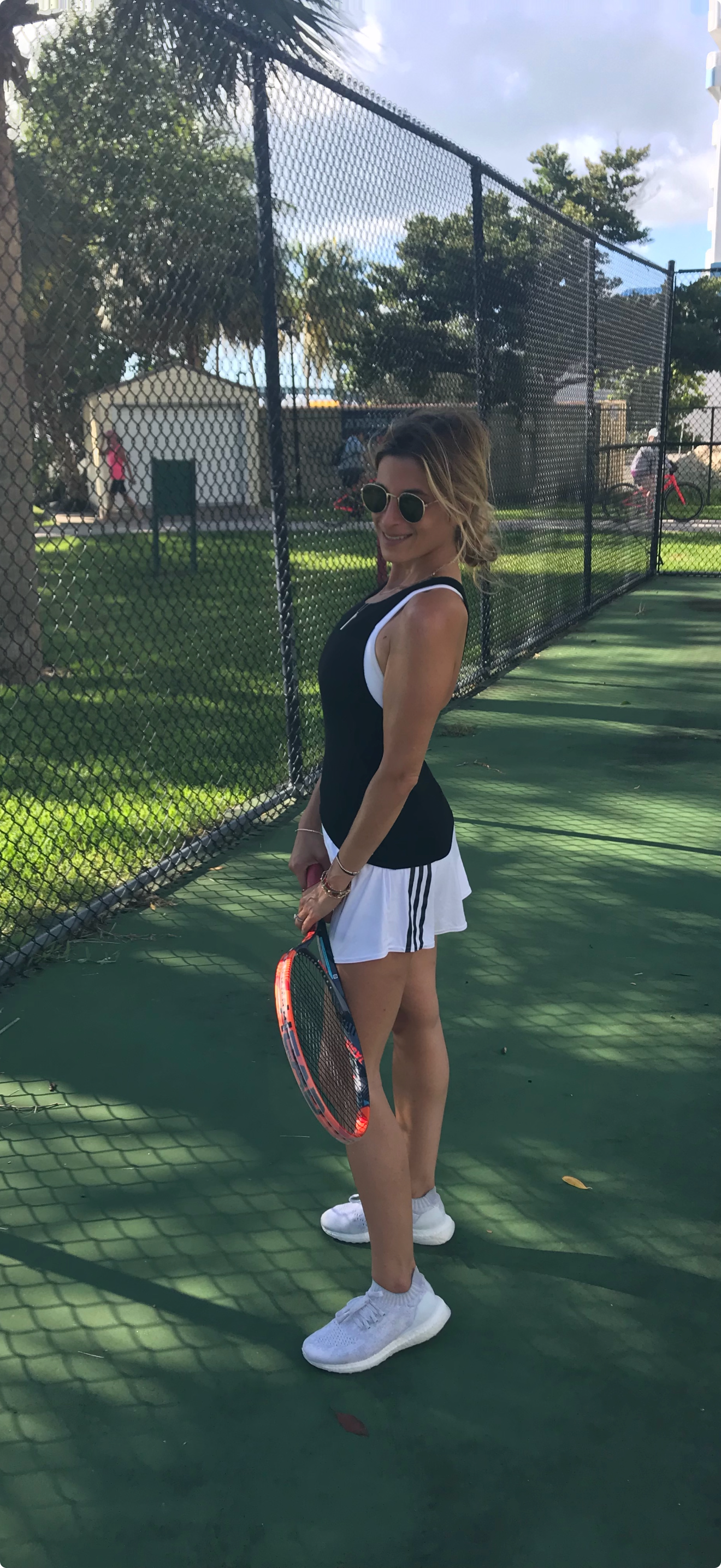 A woman in sunglasses holding a tennis racket standing on a tennis court, with a black fence and trees in the background.