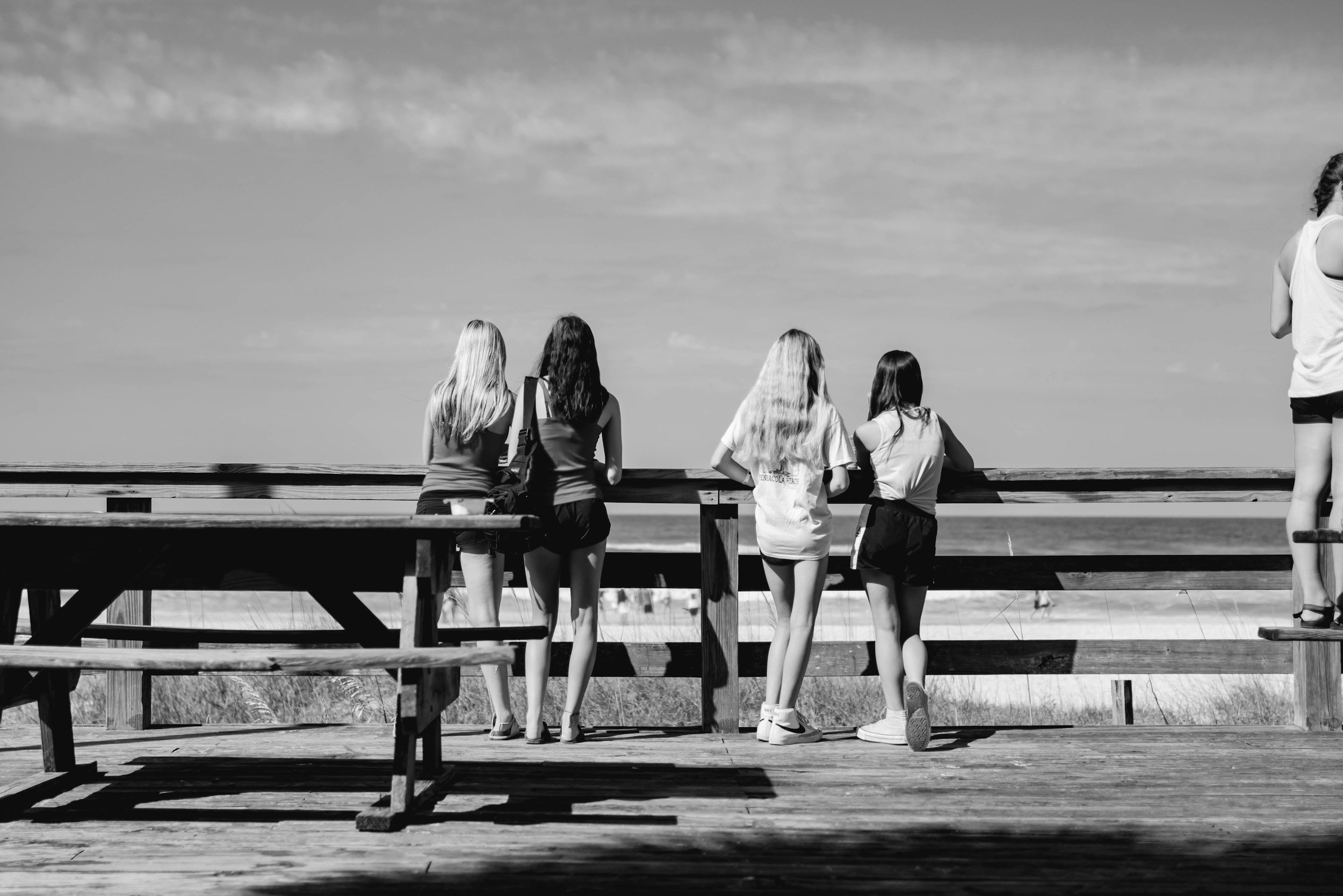 Four girls stand on a wooden pier, looking out over the ocean on a sunny day, with a fifth girl standing on a platform to the right.