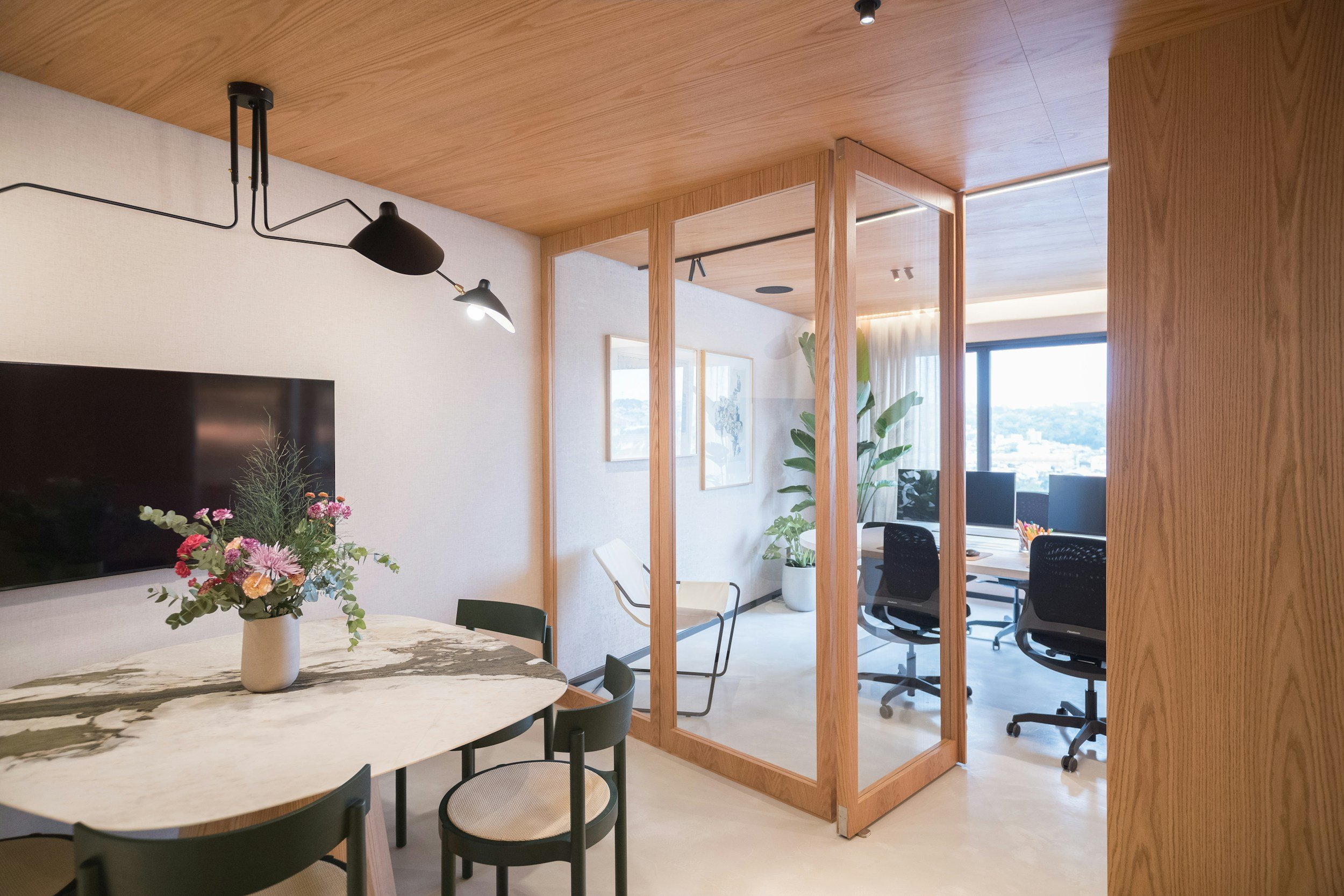 Office space with a dining area featuring a marble table, black chairs, and a flower arrangement. A glass partition separates the dining area from a workspace with multiple desks and computer monitors, large windows, and indoor plants.