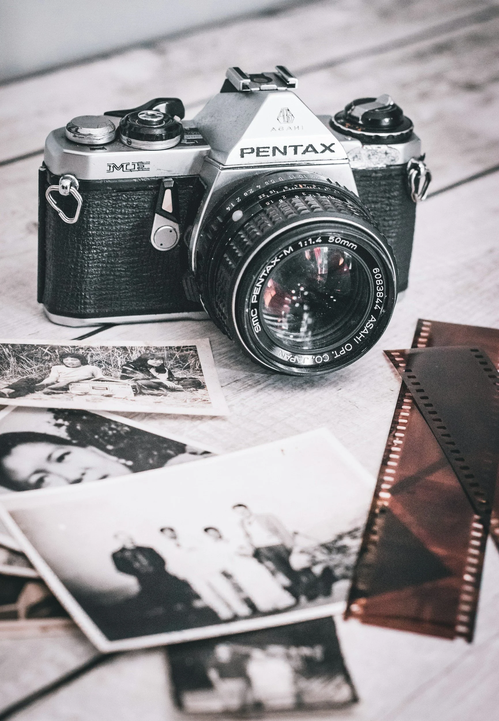 A vintage Pentax ME camera with a 50mm lens placed on a wooden surface, surrounded by black-and-white photographs and a strip of film.