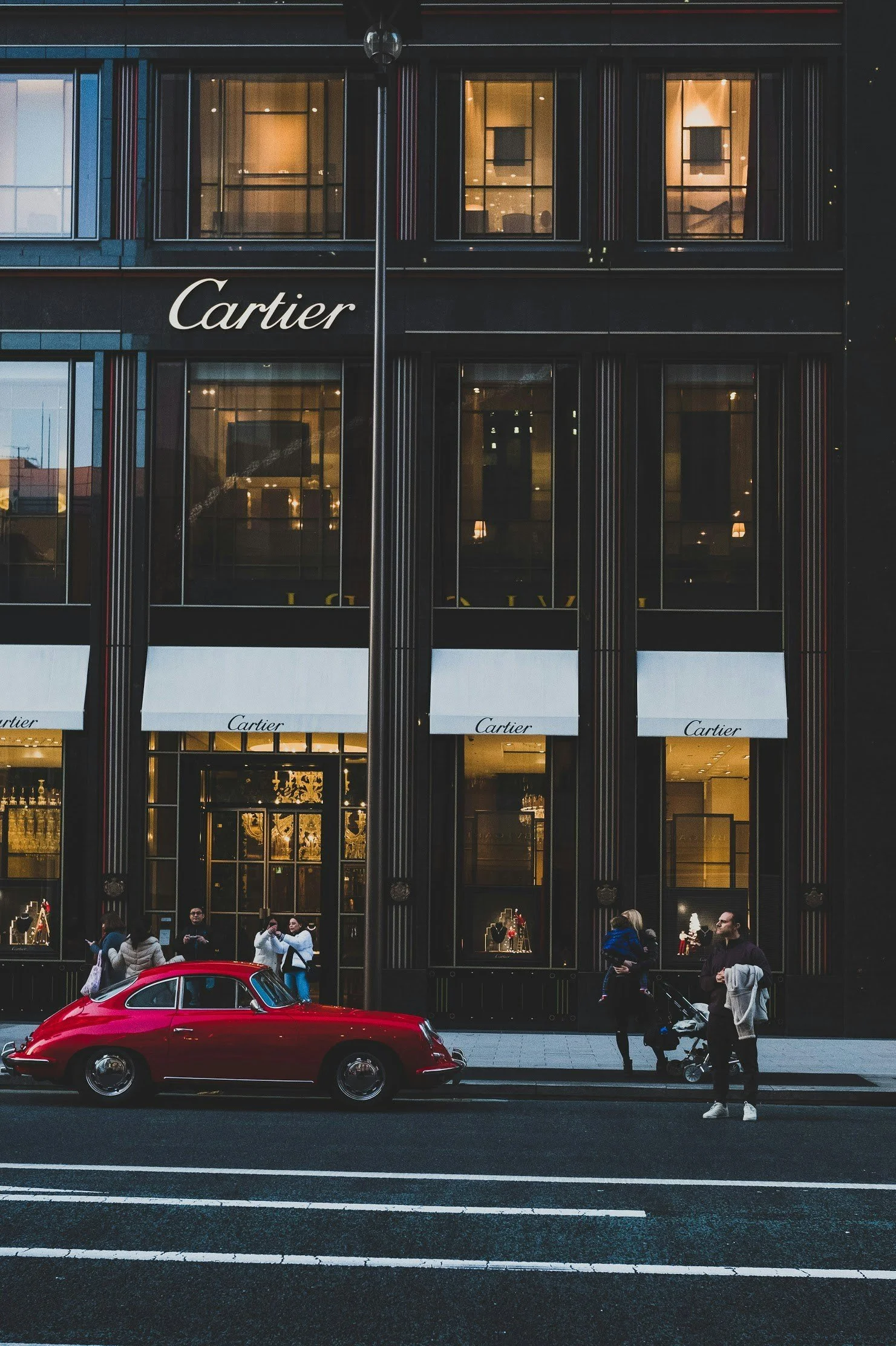 Street scene in front of a Cartier store with large windows and white awnings. A red vintage car is parked on the street, and several pedestrians are walking by and standing on the sidewalk.