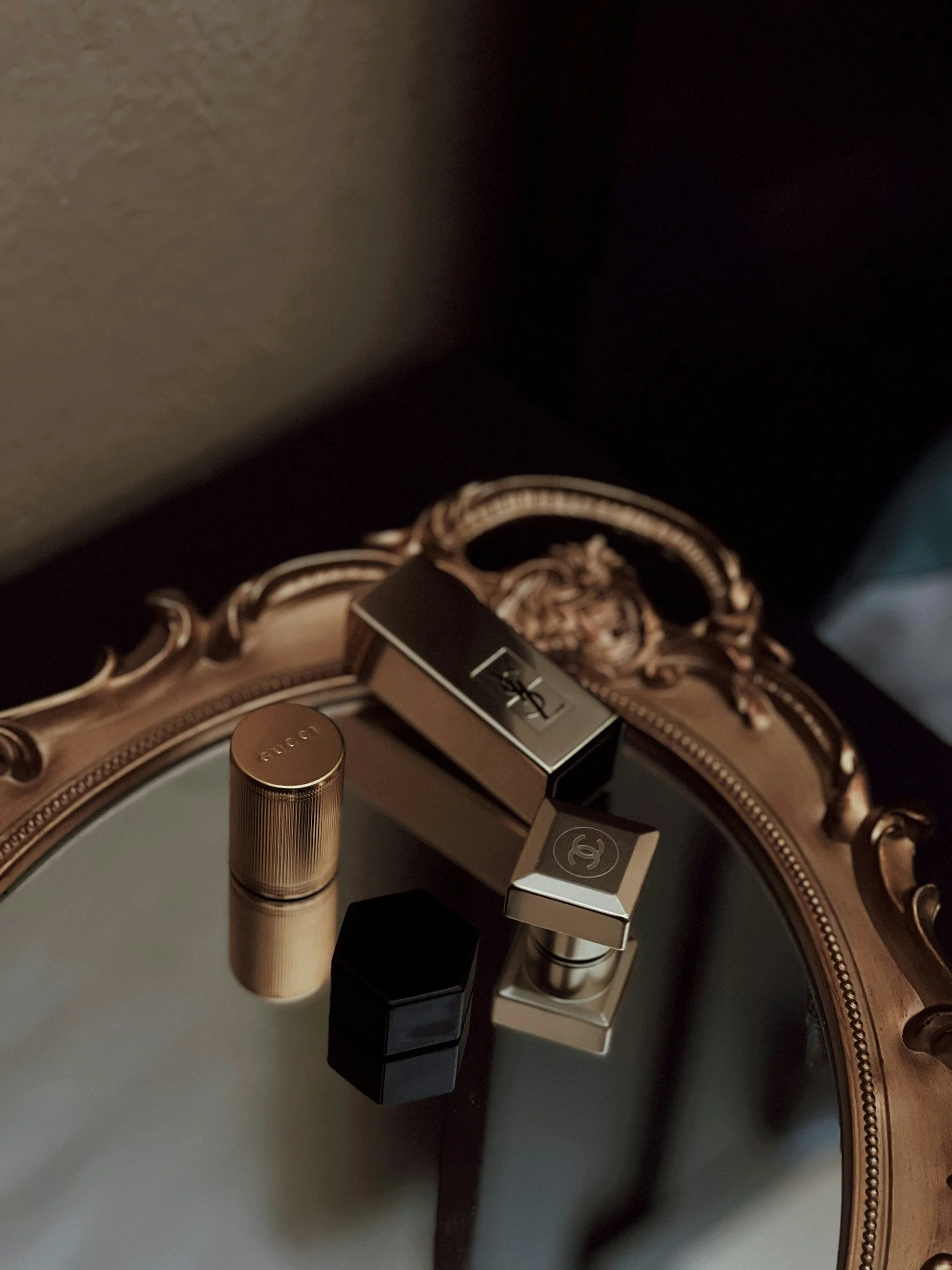 Four luxury lipstick cases with Gucci, Yves Saint Laurent, and Chanel logos reflect on a vintage ornate mirror surface.