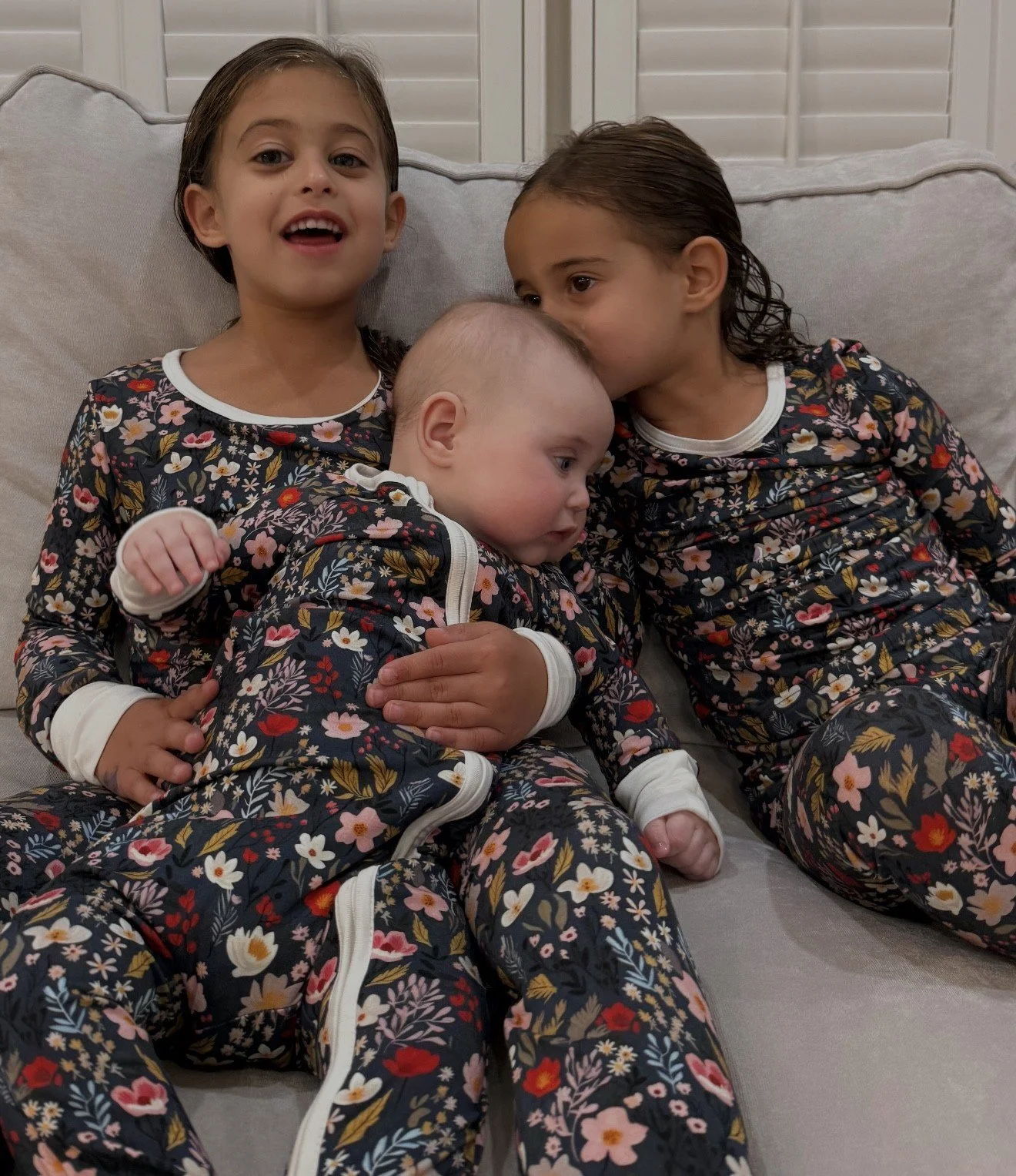 Three children sitting on a sofa, dressed in matching floral pajamas, with two older girls cuddling a younger baby.