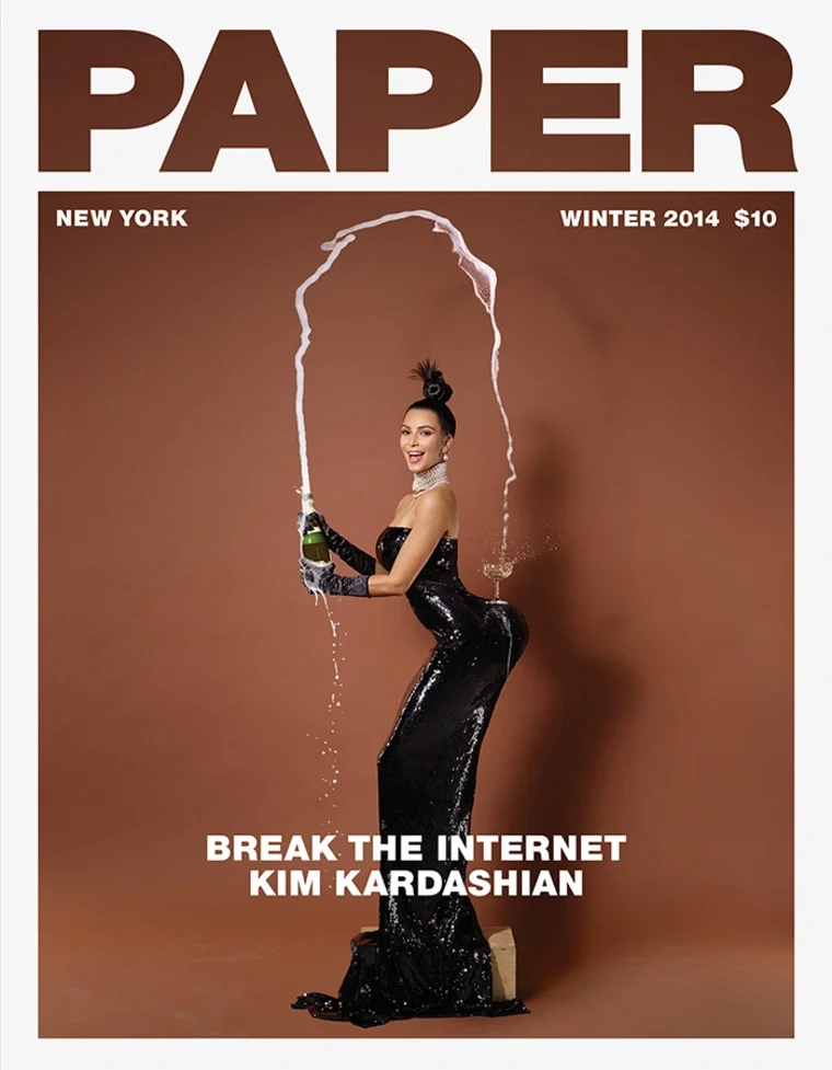 The cover of Paper magazine featuring Kim Kardashian in a black, shiny, strapless gown, holding a champagne bottle with splashes of champagne arcing in the air, against a brown background, with headlines about breaking the internet and Kim Kardashian.
