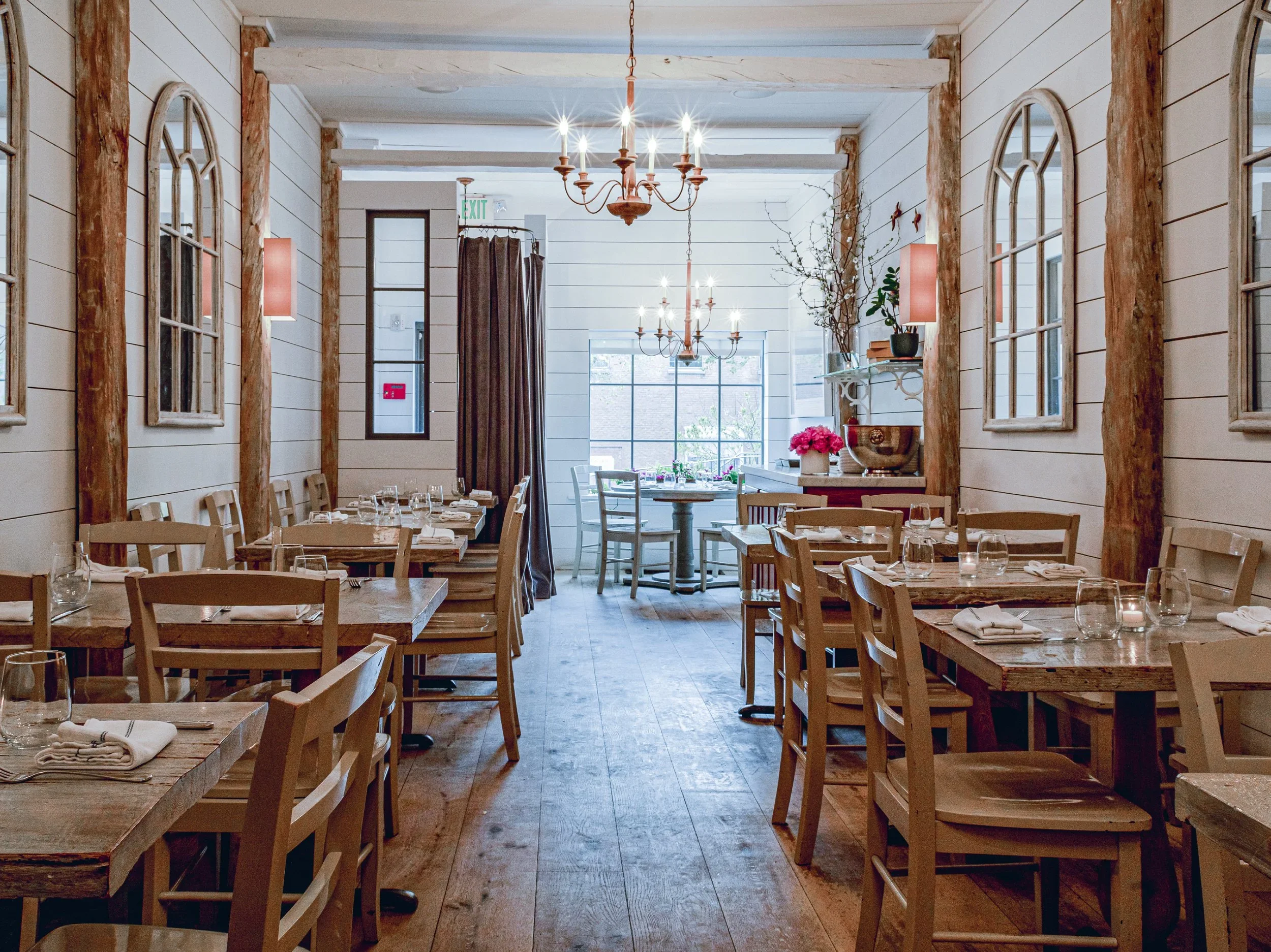 Interior of a rustic restaurant with wooden tables and chairs, chandeliers, and large windows with arched frames, decorated with plants and flowers.