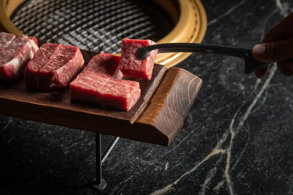 Raw beef steaks on a wooden cutting board, with a hand holding tongs. Grill in the background.