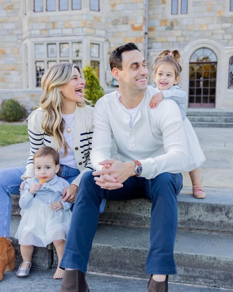 Family of four sitting on steps in front of a stone building, smiling and enjoying each other's company.