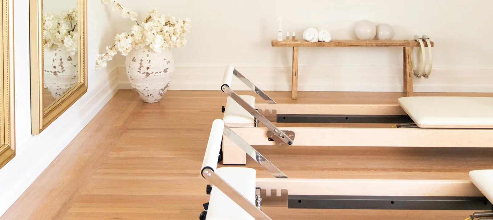 Reformer Pilates machine in a serene, well-lit room with wooden flooring and white walls, decorated with large flower vase, mirror, and minimalist decor.