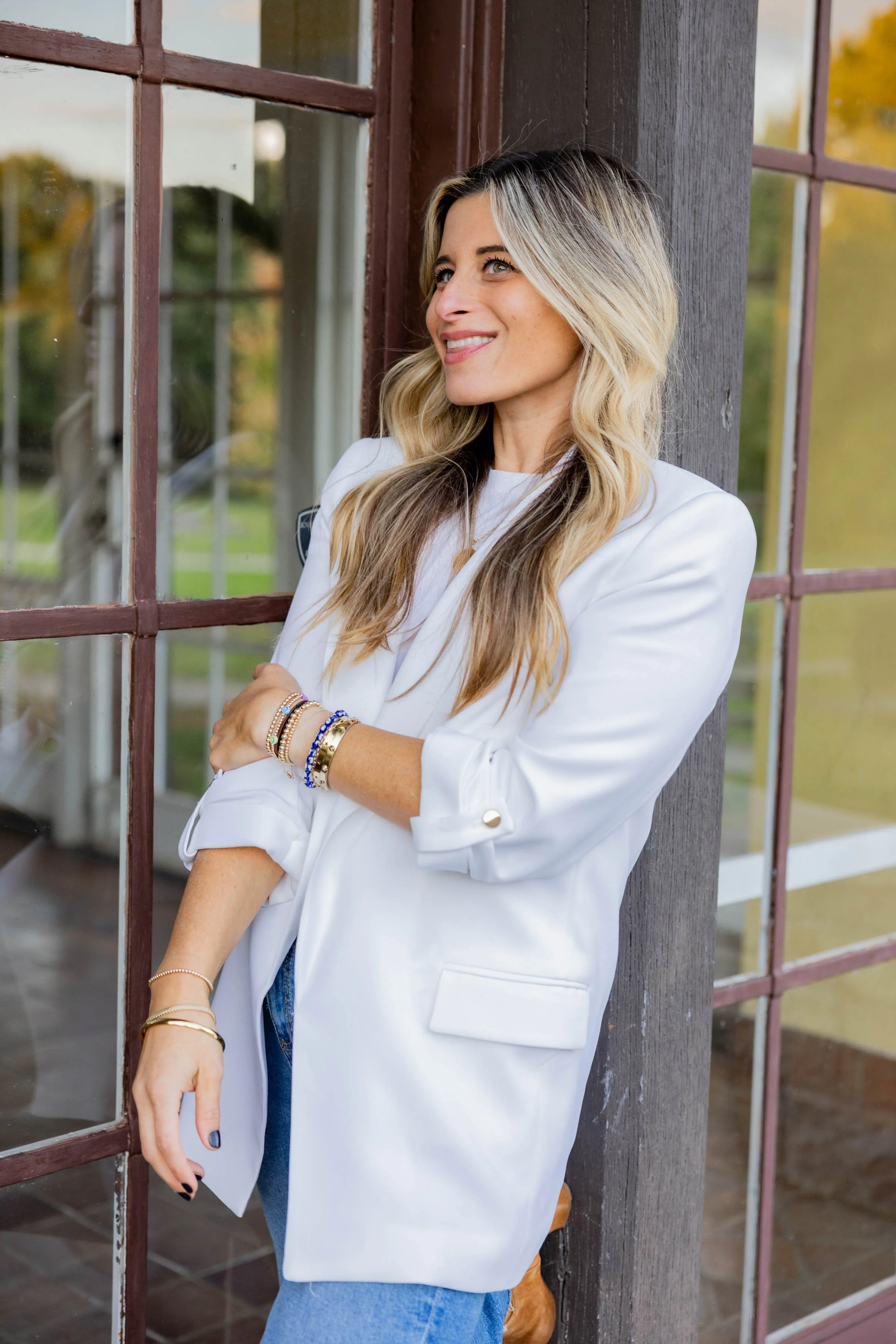 Carrie Georgette Kerner A smiling woman with long blonde hair, wearing a white blazer and blue jeans, leaning against a wooden beam near large glass windows with an outdoor view of trees and greenery.