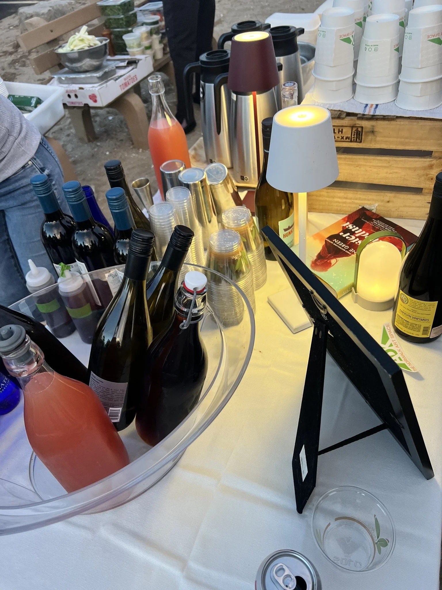A beverage stand with bottles of wine and soda, glasses, paper cups, and lamps on a table, likely at an outdoor event or market.