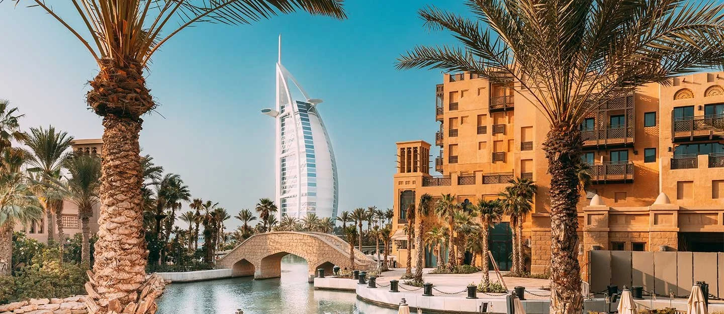 View of the Burj Al Arab hotel with palm trees, a waterway, and a bridge at sunset.