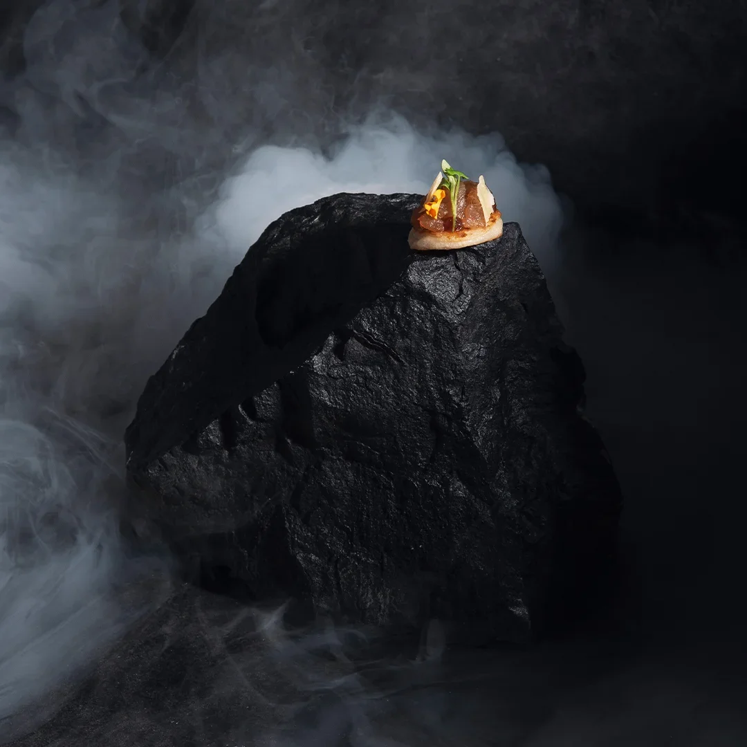 A piece of sushi with fish, green garnish, and yellow flower on a large black rock surrounded by white mist.