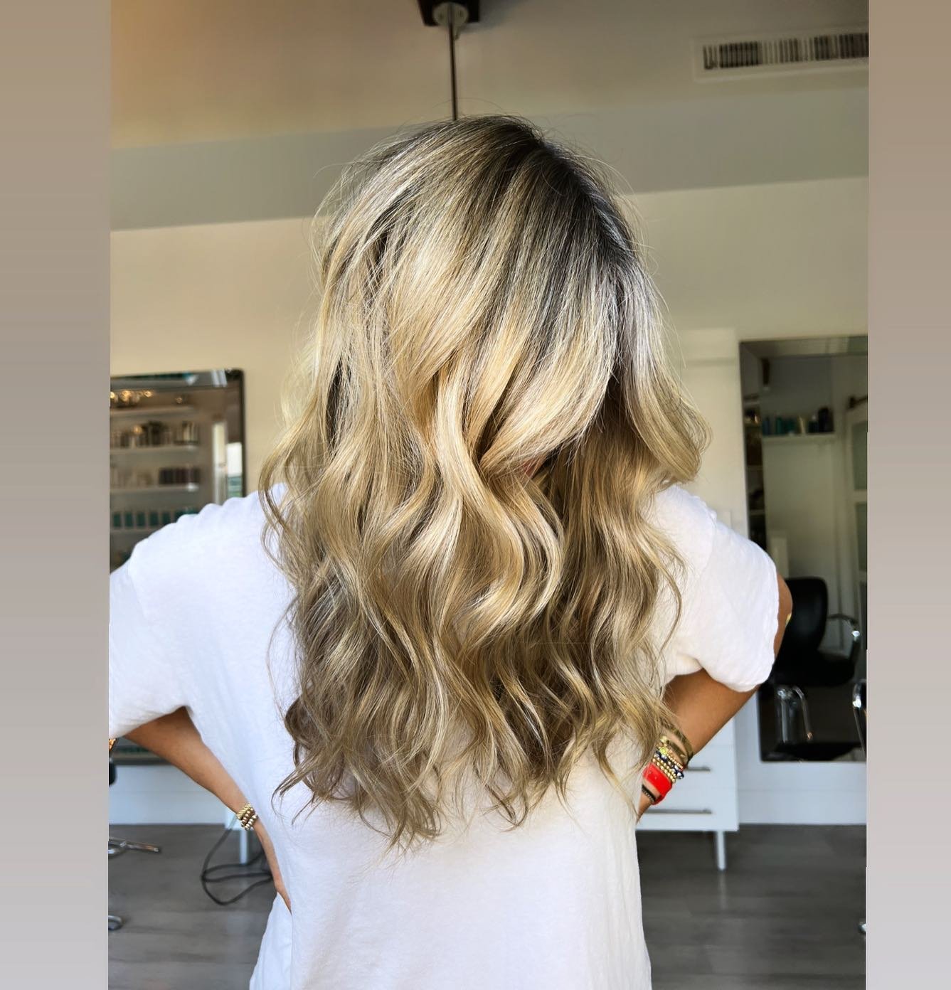 Back of a Carrie Georgette Kerner  with long, wavy blonde hair standing indoors, wearing a white top and colorful bracelets on her wrist.