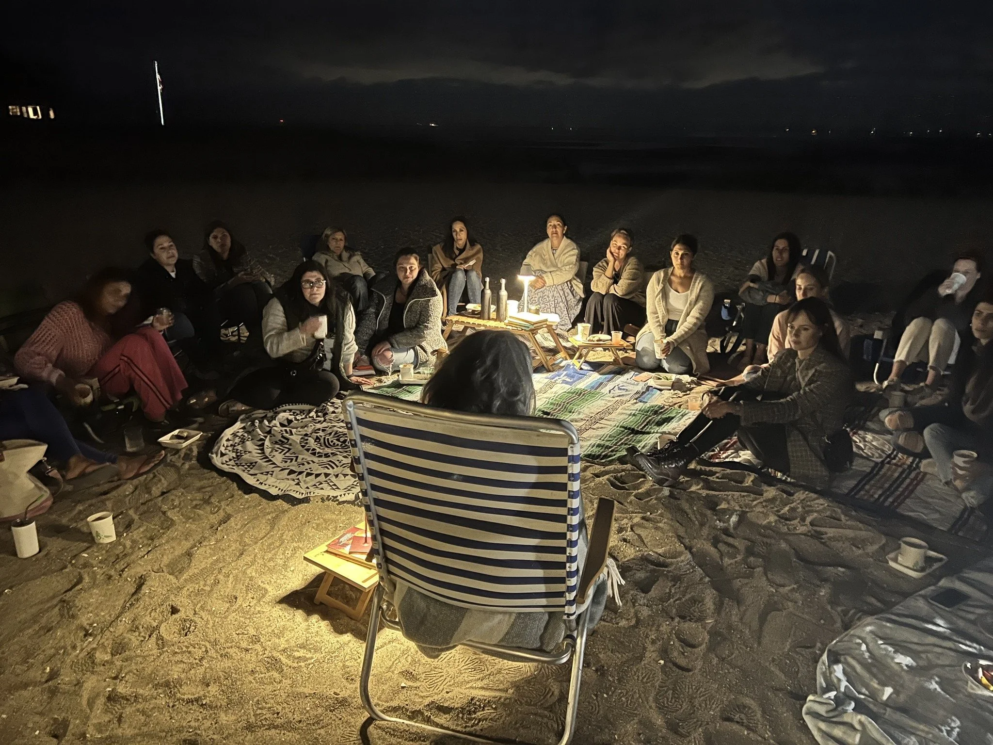 A group of people sitting on the sand around a small lamp on the beach at night, engaged in a gathering or discussion.