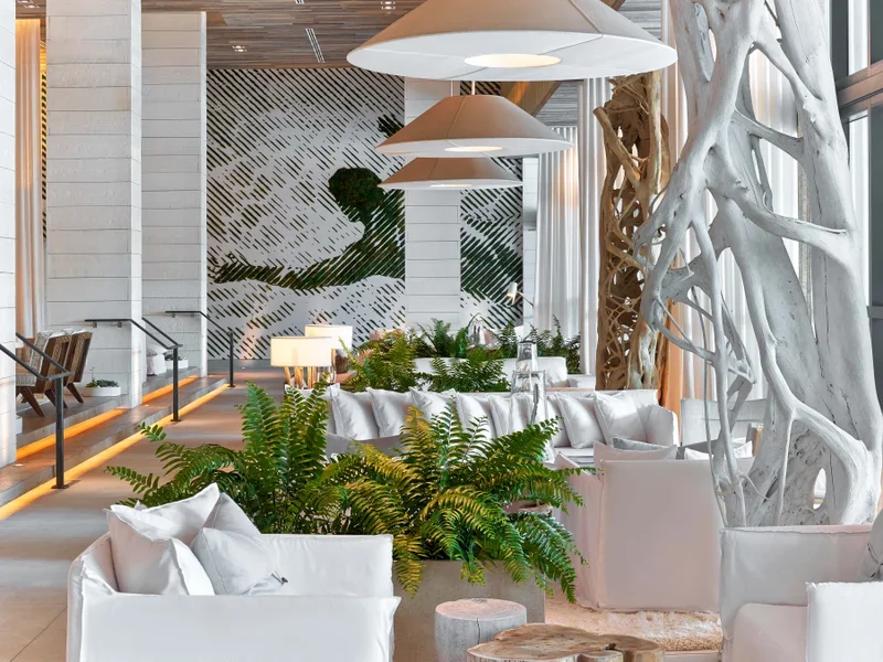 Modern indoor lounge area with white sofas, green plants, large white decorative tree, and pendant lights.