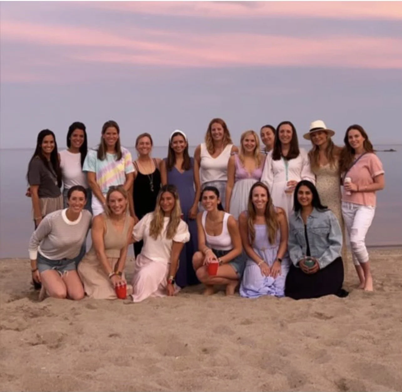 A group of 16 women posing on the beach at sunset, some holding drinks and wearing casual summer clothing. Books on the Beach 203 Westport CT